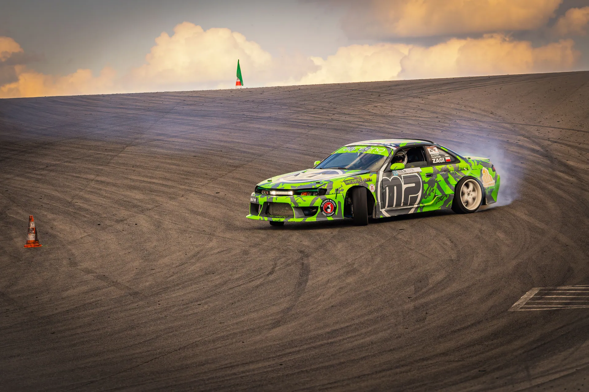 Green car drifting on a track, showcasing skill and speed against a sunset backdrop.