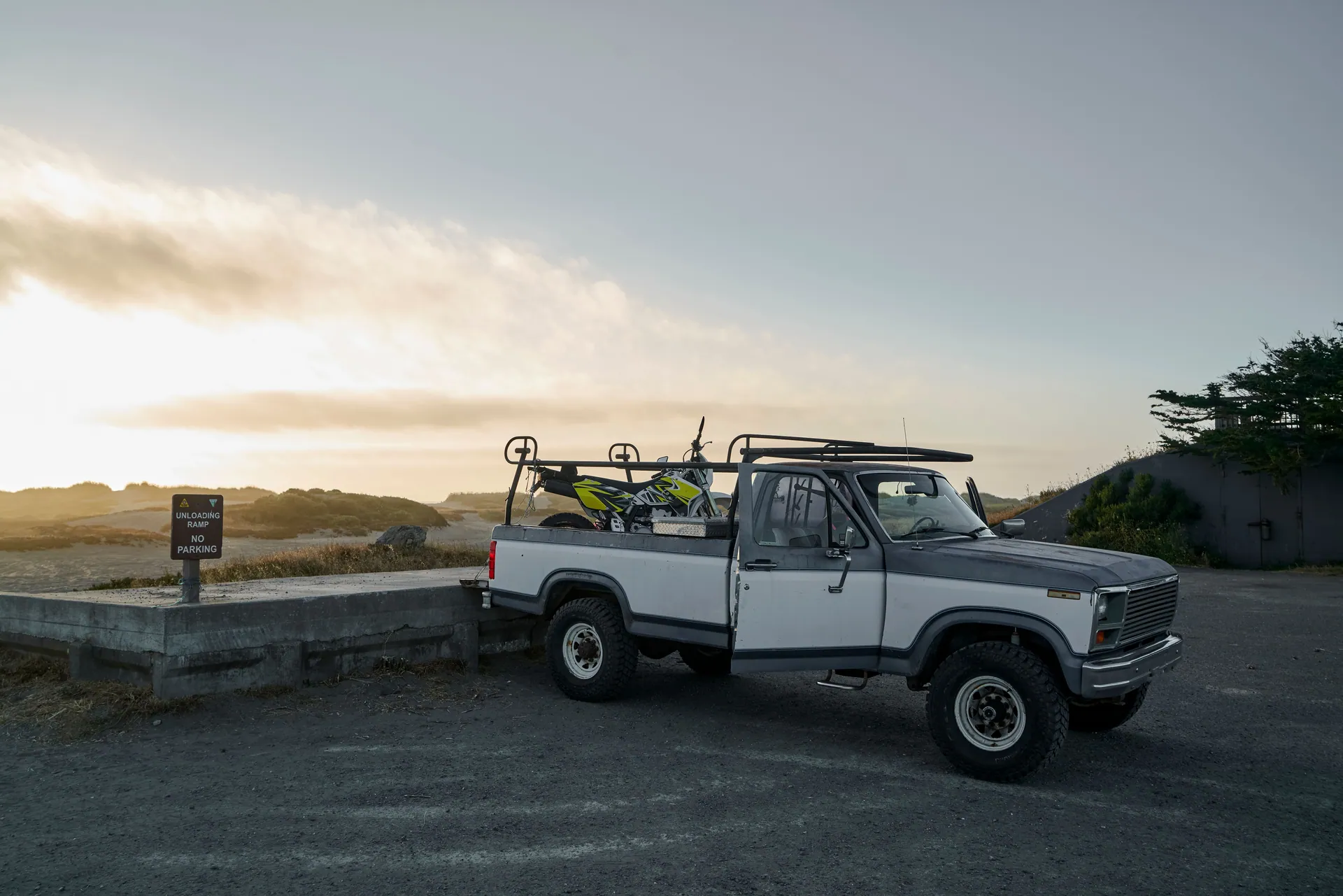 A classic pickup truck parked at a scenic spot during twilight with a dirt bike loaded on the back.