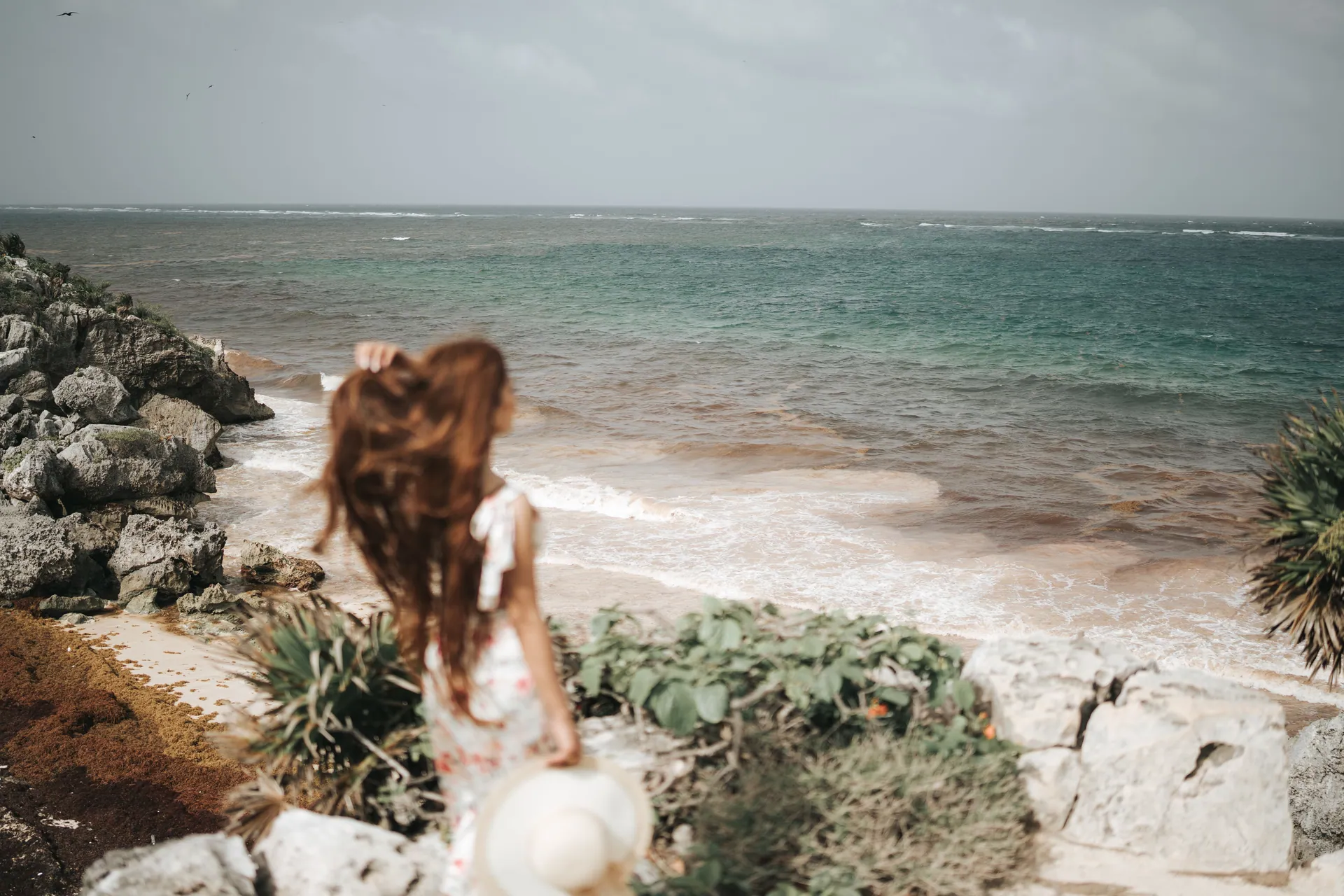 A woman with long hair stands by a rocky seashore, holding a hat in a serene coastal scene.