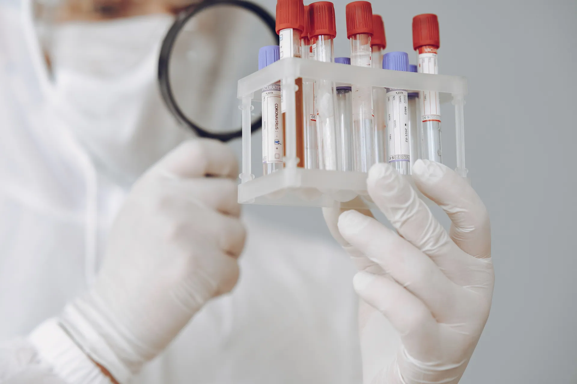 A laboratory scientist examines blood samples using protective gear and a magnifying glass.