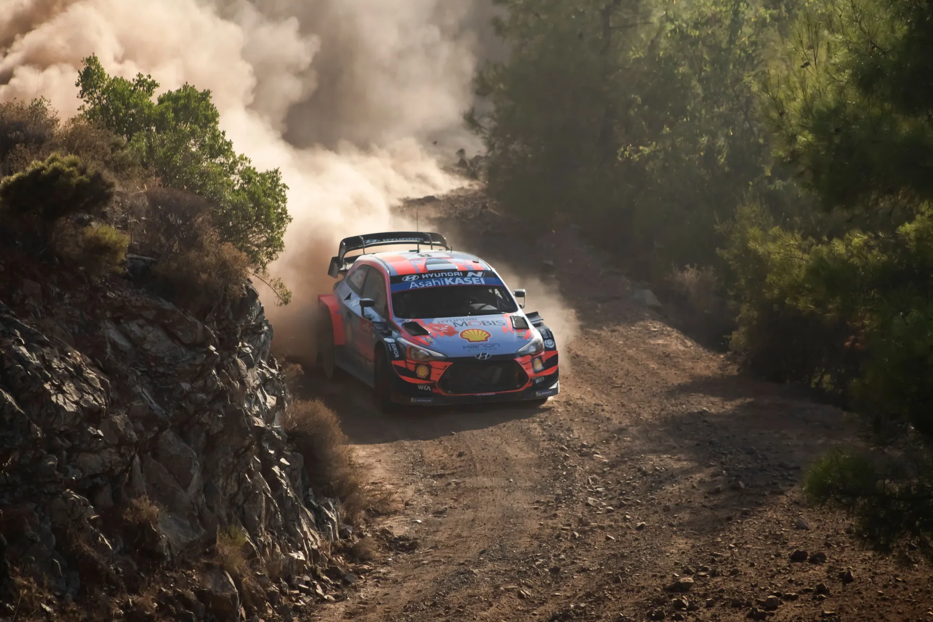 An exhilarating dirt track rally car speeds through a forested race course, casting clouds of dust.