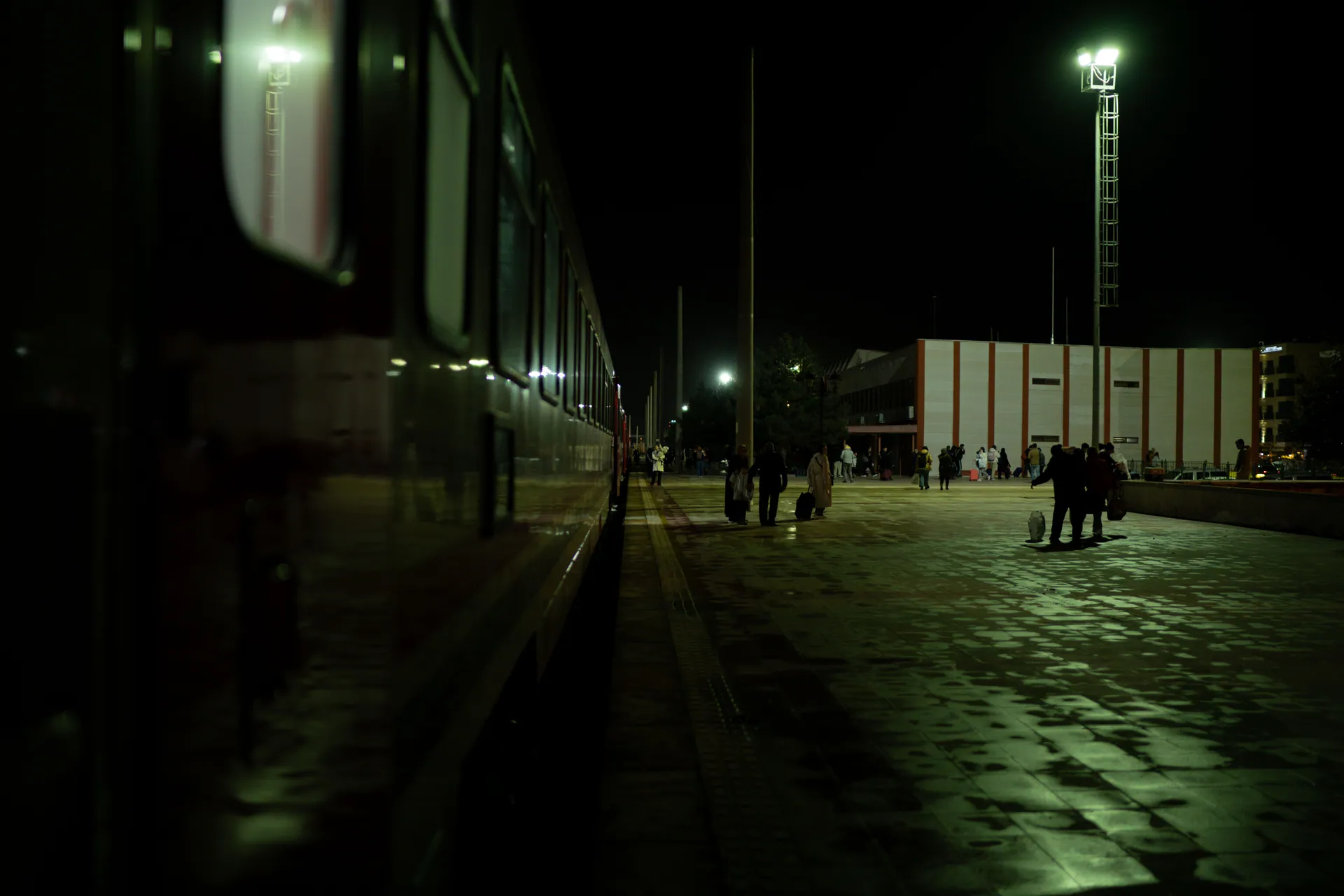A bustling train station at night with passengers walking along the platform under bright lights.