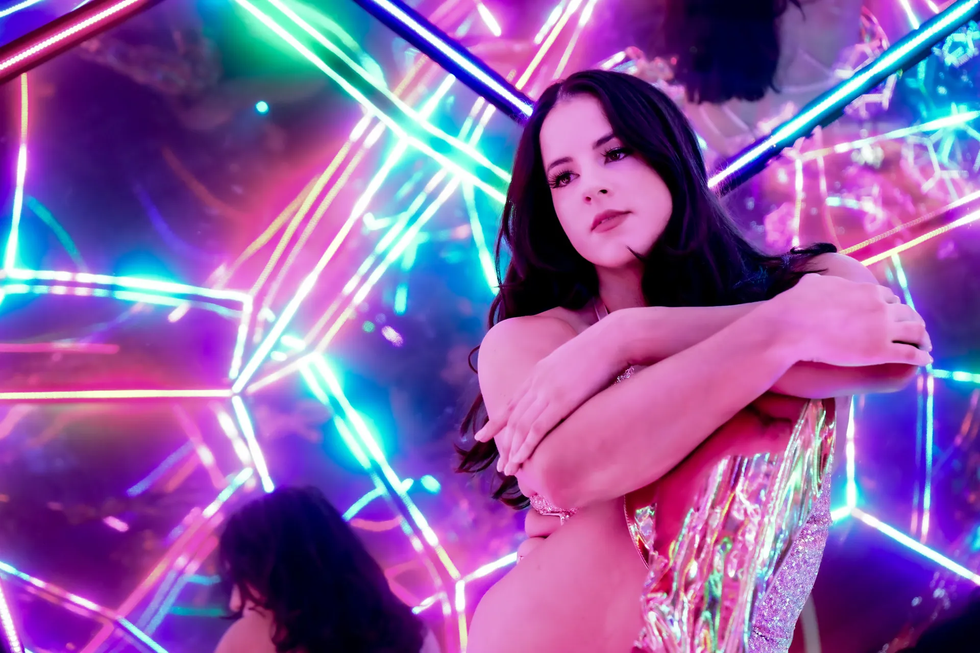A young woman poses amidst colorful neon lights, creating vibrant reflections.