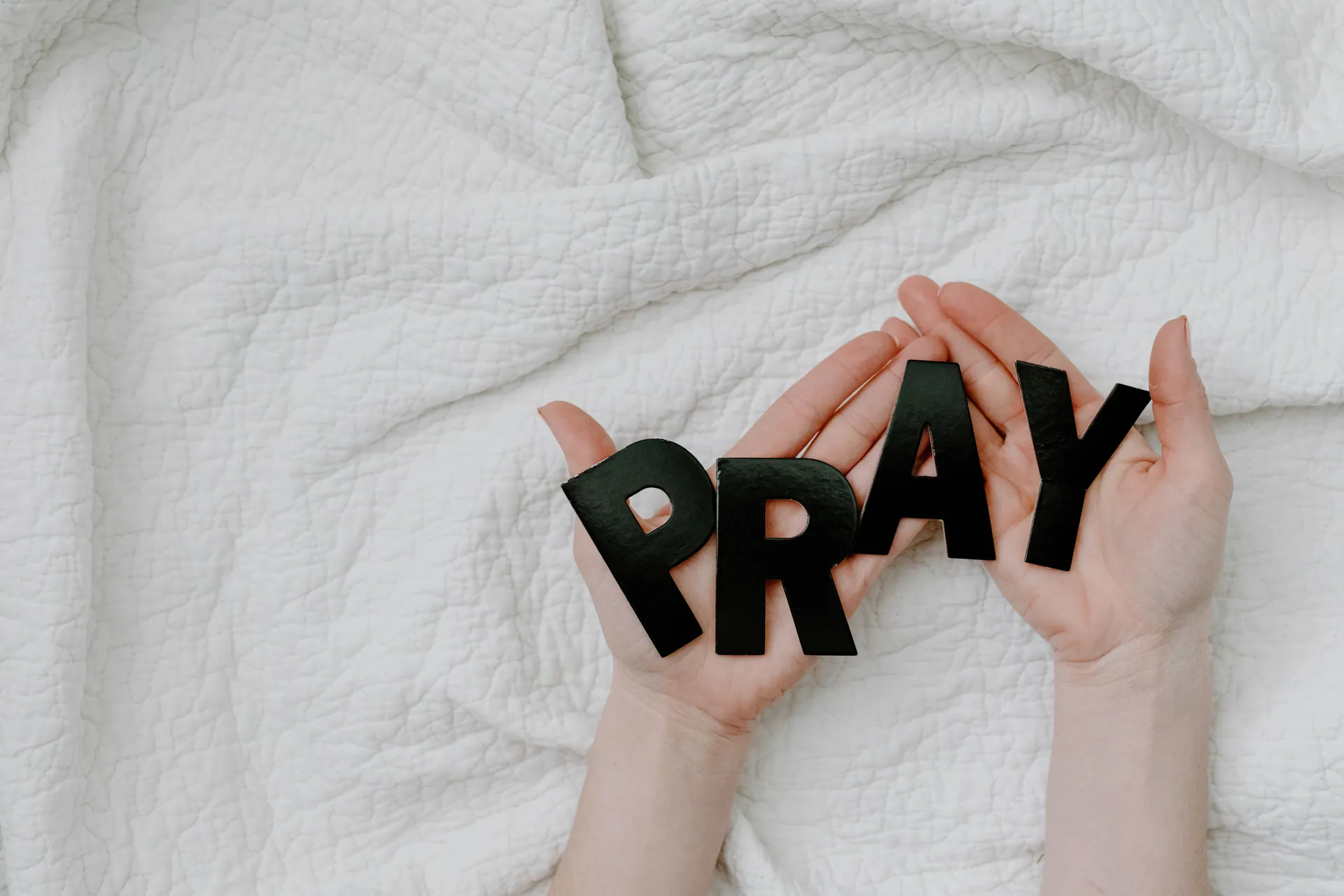 Close-up of hands holding black pray letters on white quilt background for a prayer theme.