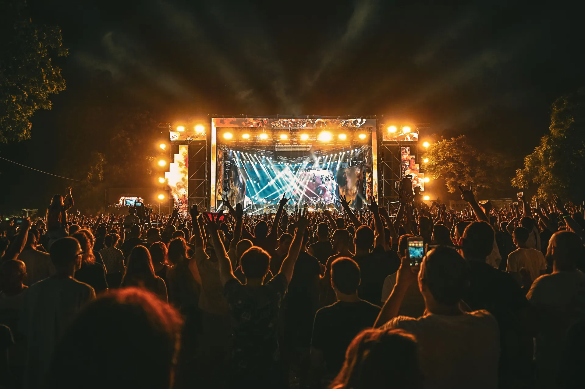 Large crowd enjoying a spectacular live outdoor concert at night with stage lighting effects.