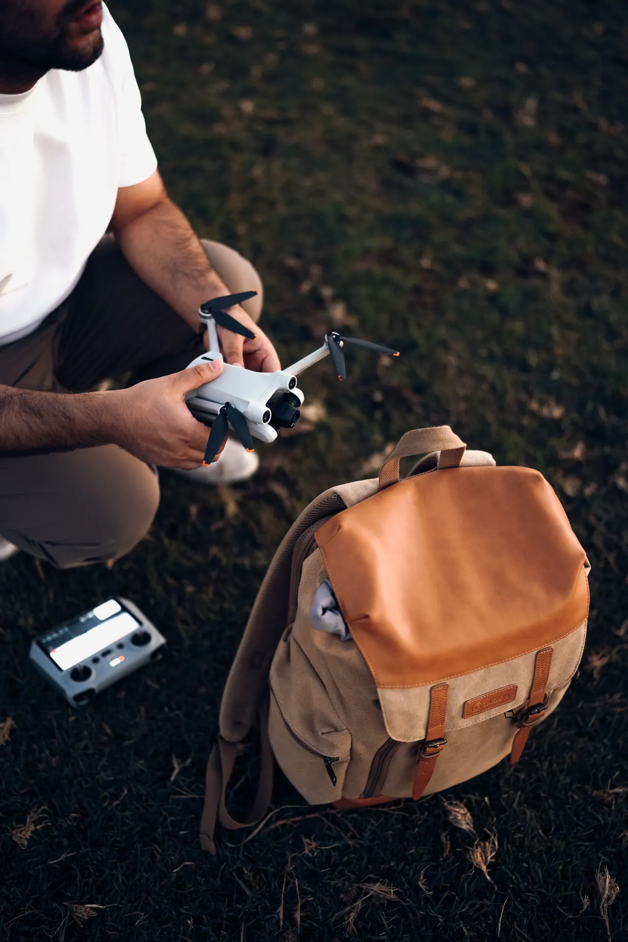 A man crouches with a drone and backpack, ready for an outdoor adventure.