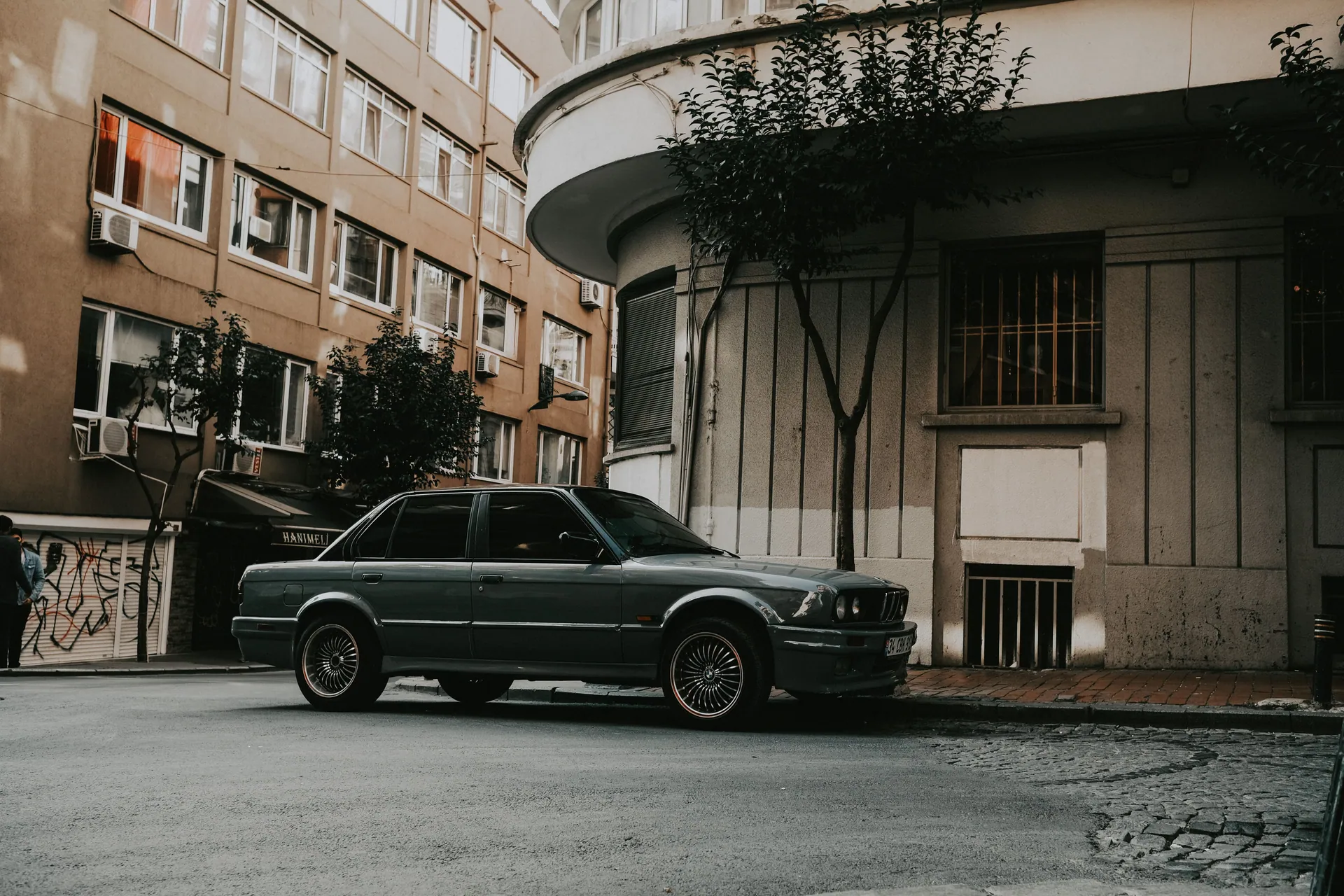 A classic car is parked on a city street, surrounded by urban architecture and trees.