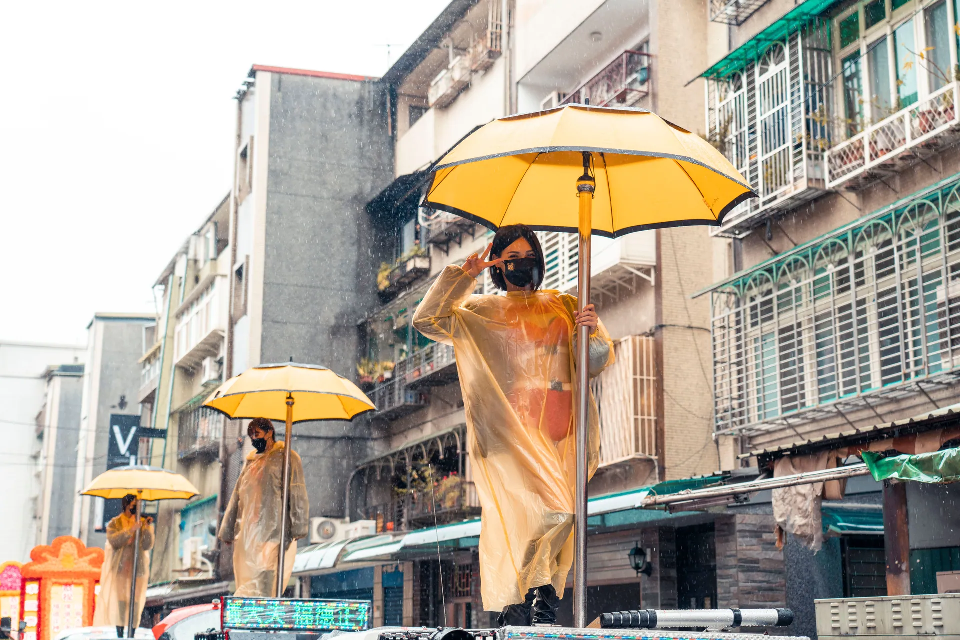 Women in raincoats and face masks hold yellow umbrellas during a rainy day parade in an urban setting.