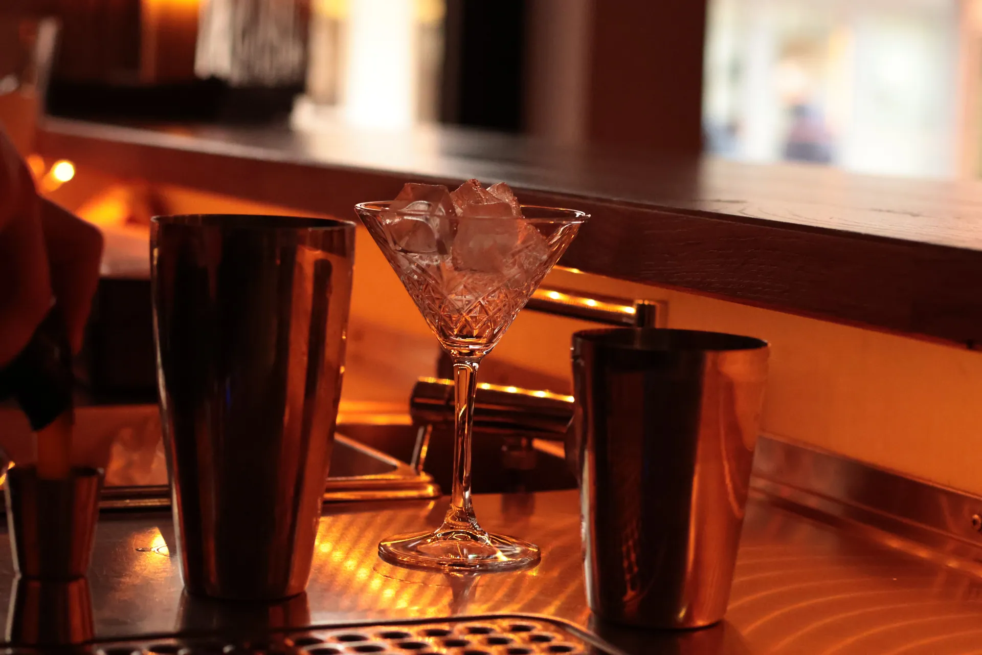 A stylish bar counter features a martini glass with ice, surrounded by cocktail shakers in a warm ambiance.