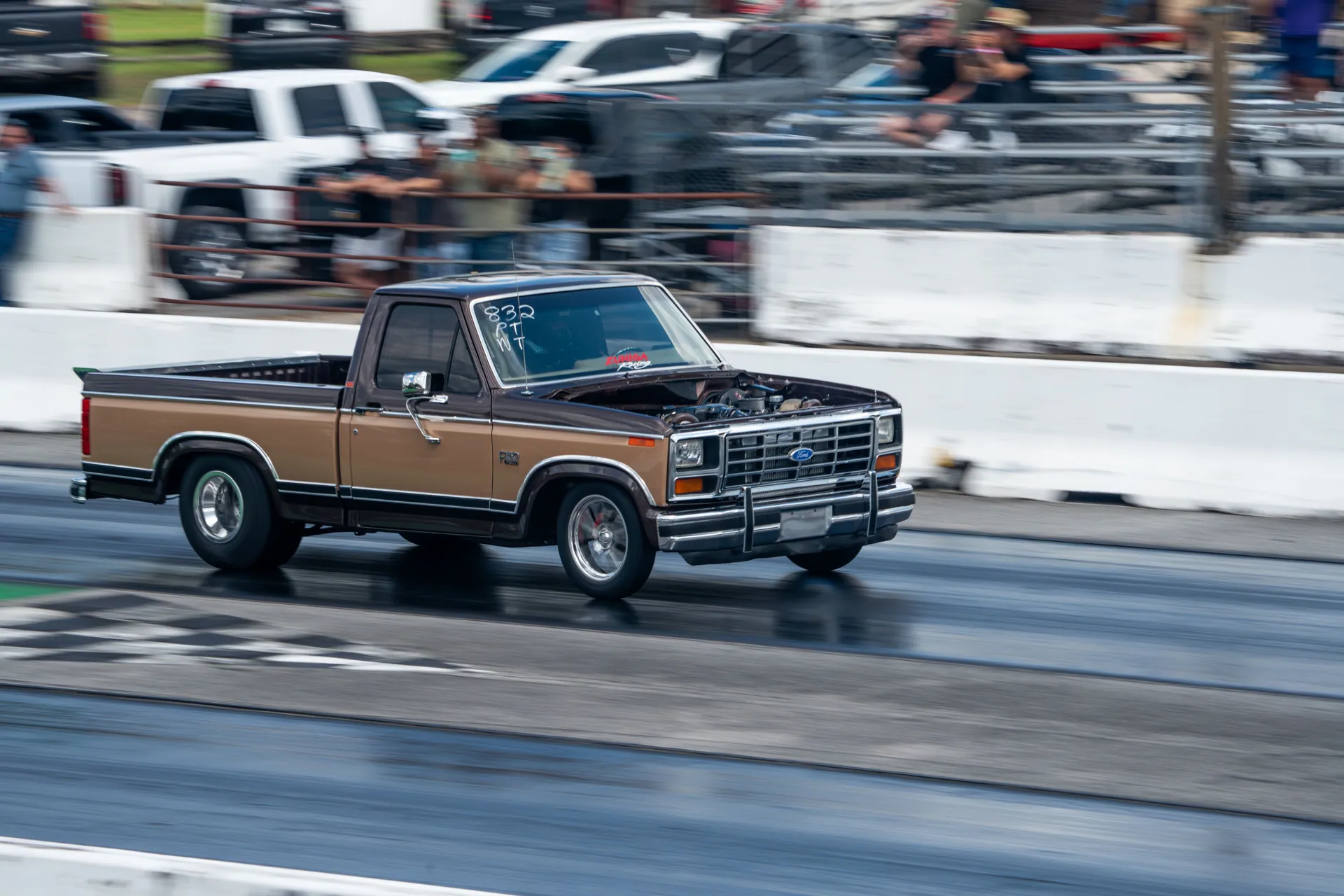 Vintage brown pickup truck speeding on drag strip, captured in motion at daytime.
