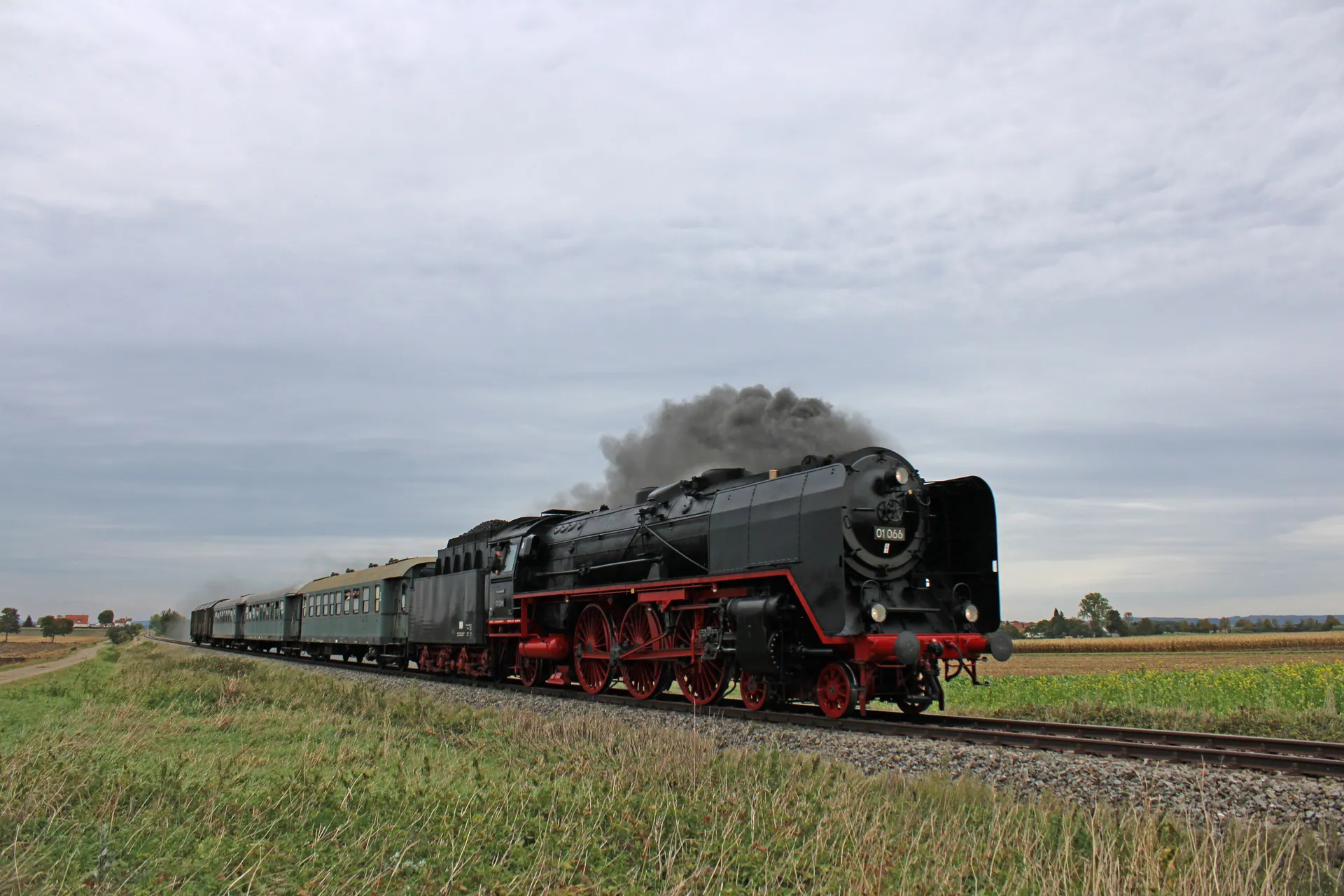 A vintage steam train travels through rural fields under a cloudy sky, showcasing classic transportation.
