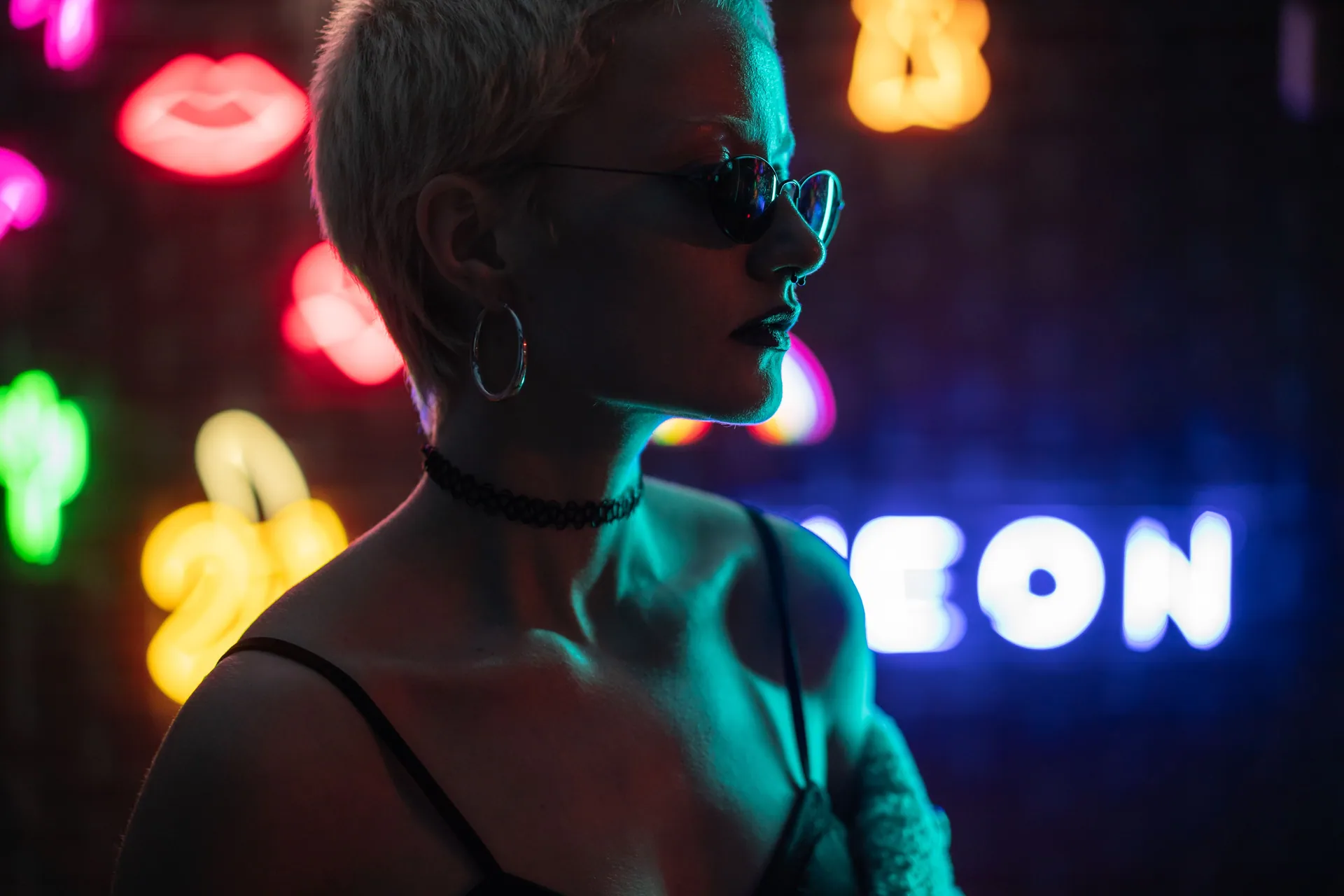 Stylish woman in sunglasses posing against colorful neon lights at night.