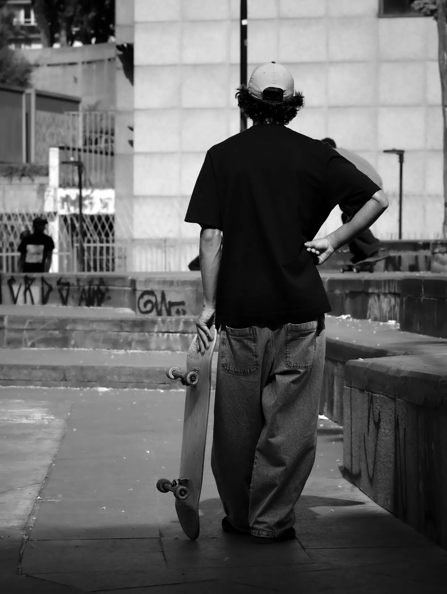 A skateboarder stands with his board in a gritty urban setting, captured in black and white.