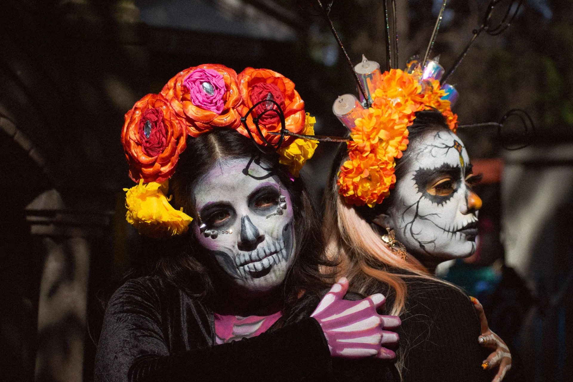 Two people in colorful Day of the Dead makeup and floral headpieces embracing outdoors.