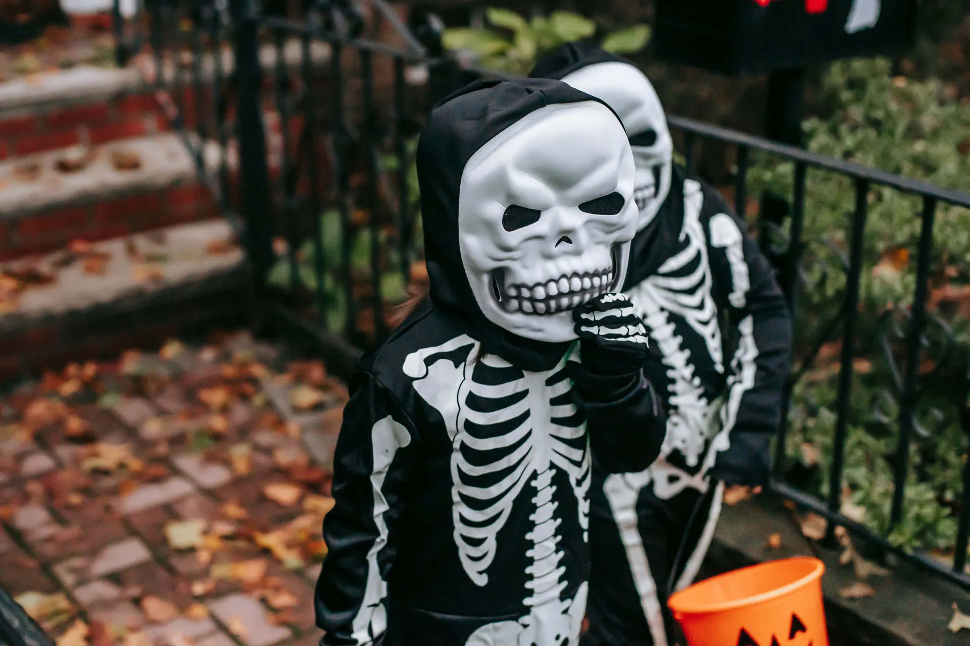Faceless kids in black skeleton costumes with masks and orange bucket trick or treating in Halloween in daytime