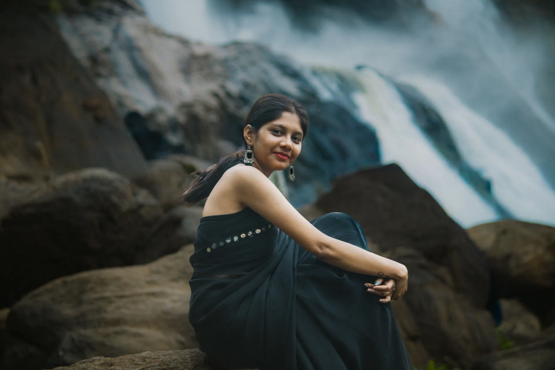A woman in a black sari seated by rocks near a scenic waterfall in Pariyaram, India.