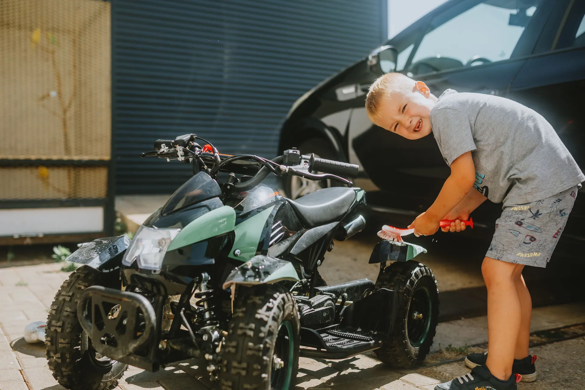 A young boy cheerfully cleans a kids' ATV outdoors on a sunny day, showcasing a playful moment.