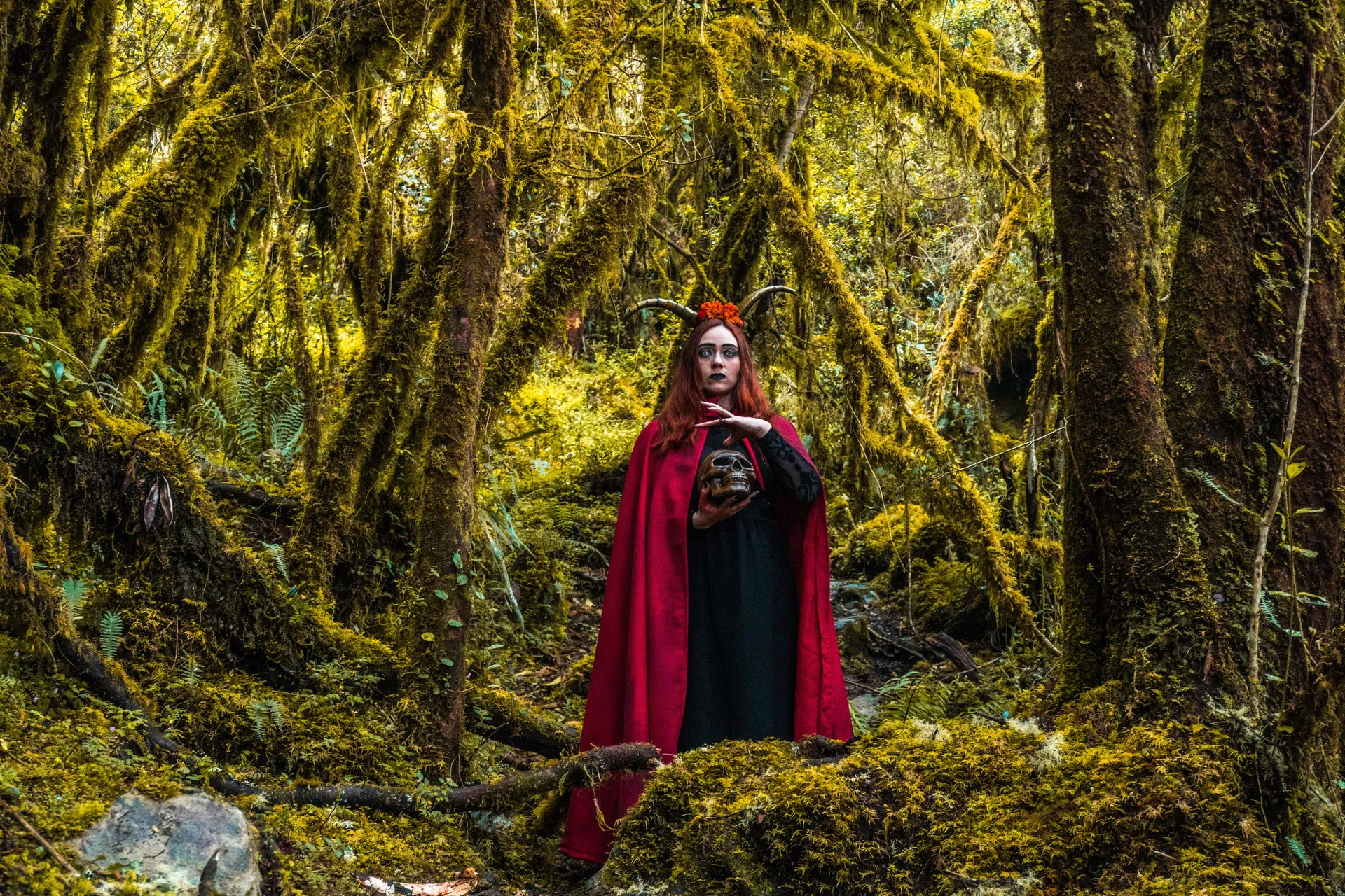 A woman in a red cloak with horns stands in a lush, mossy forest holding a skull, embodying a mystical and spooky Halloween scene.