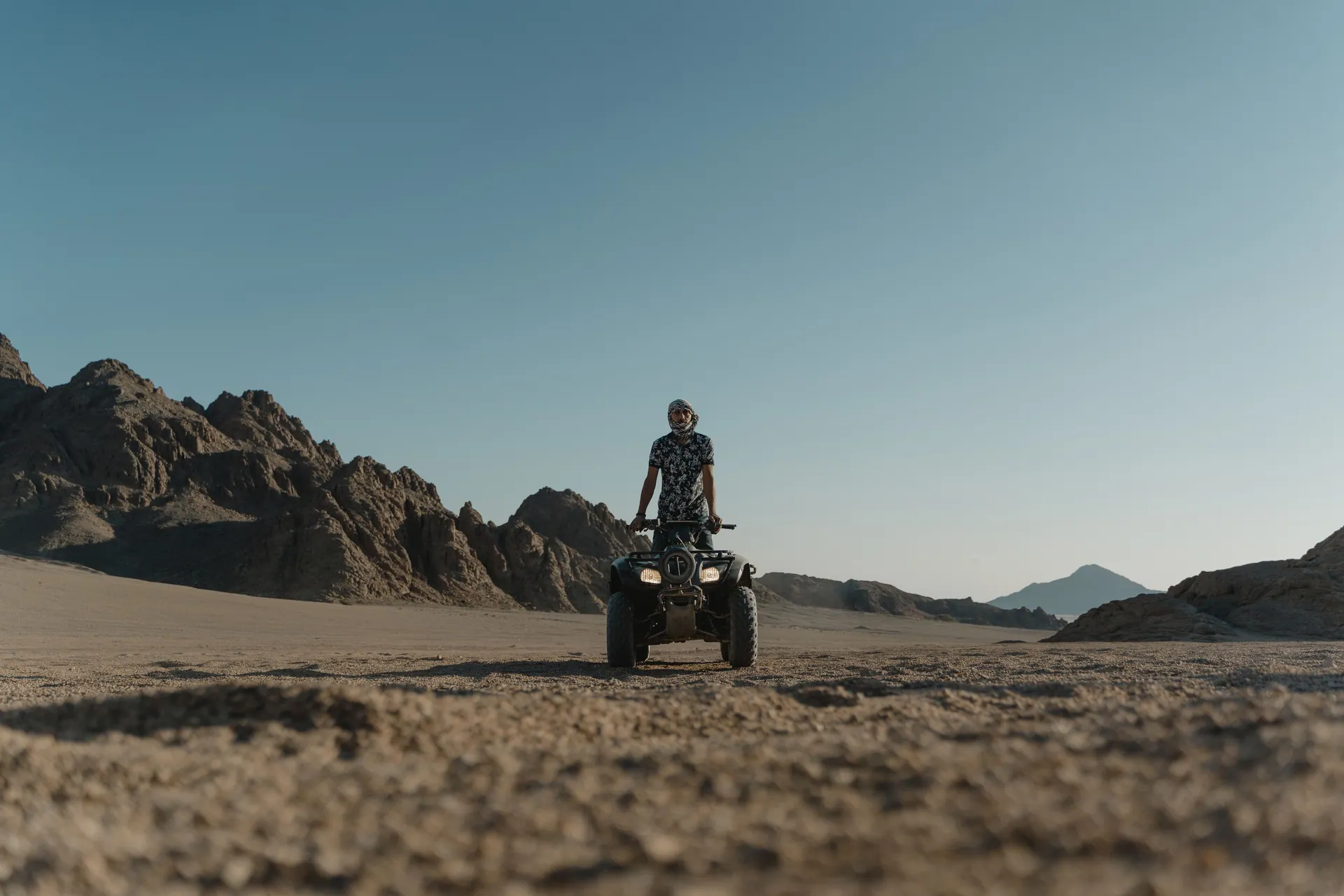 Person standing on a quad bike in a sandy desert with rocky mountains in the background.