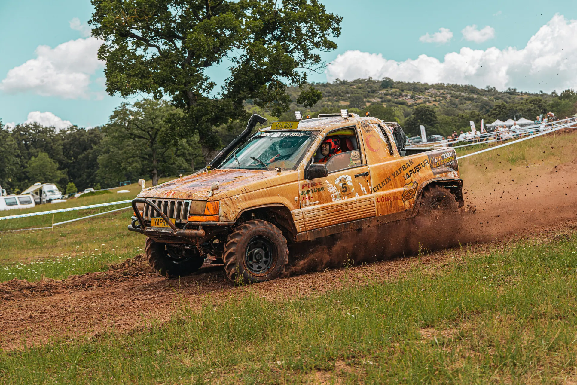 Thrilling offroad vehicle race on a muddy track amidst a lush countryside landscape under a bright sky.