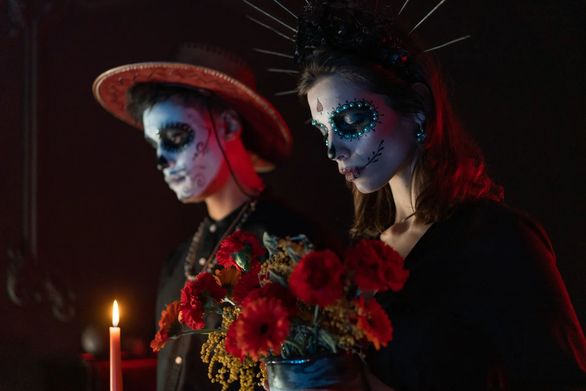 Couple celebrating Dia de los Muertos with traditional face paint and flowers in a dark setting.