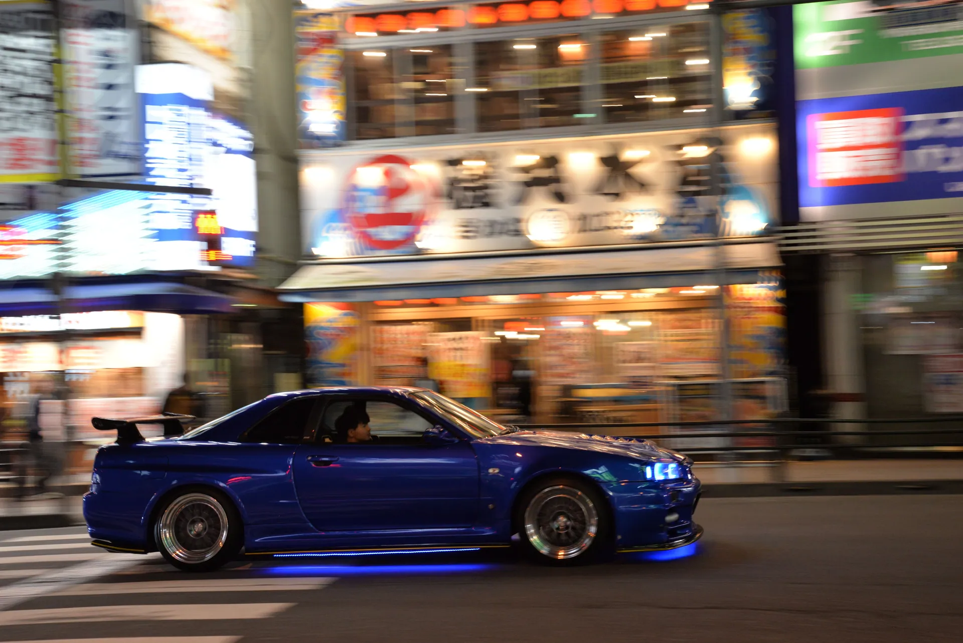 Dynamic shot of a blue sports car speeding through vibrant Tokyo streets at night.
