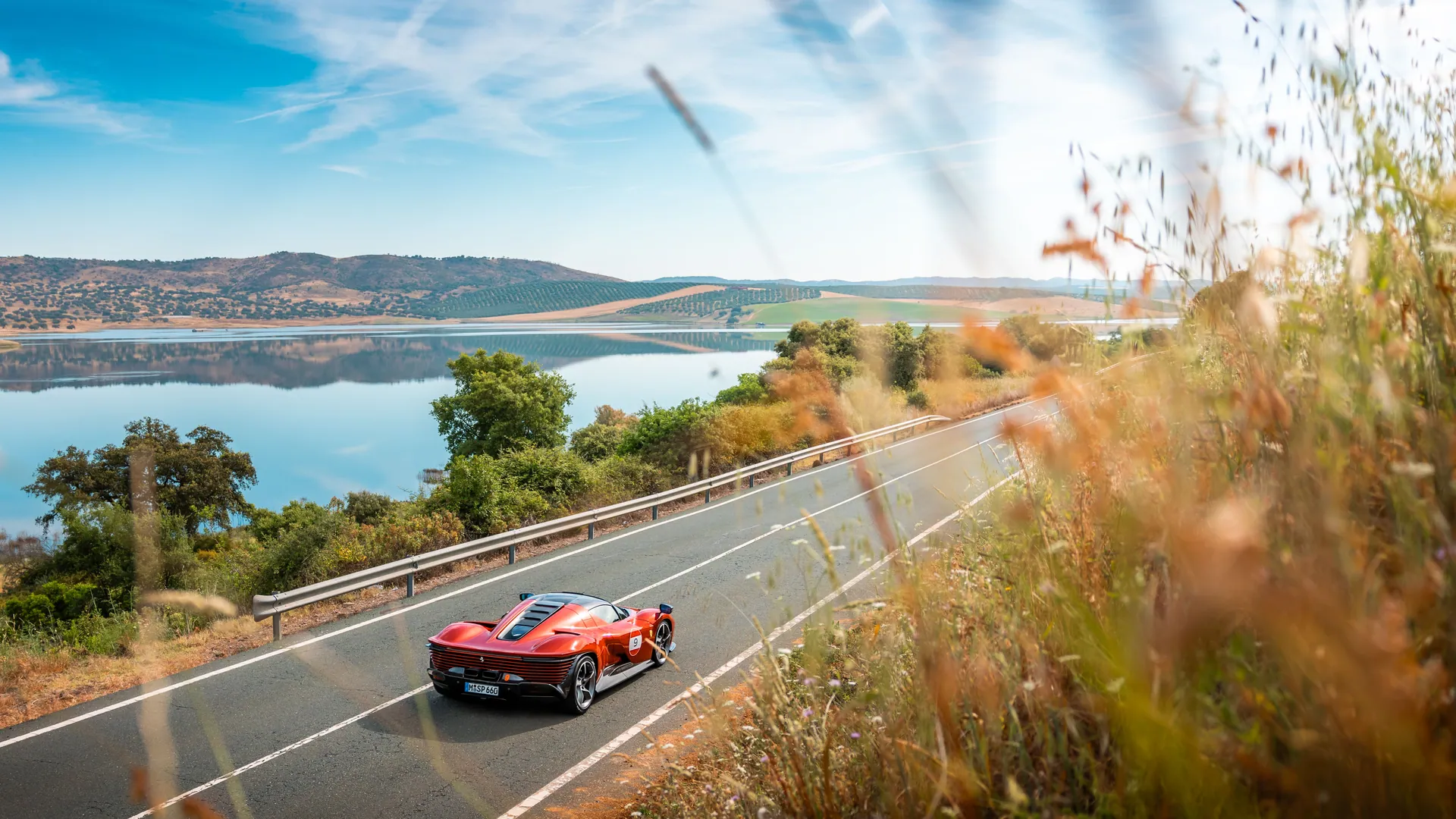 A stunning red sports car cruising on a picturesque road beside a serene lake under a clear blue sky.