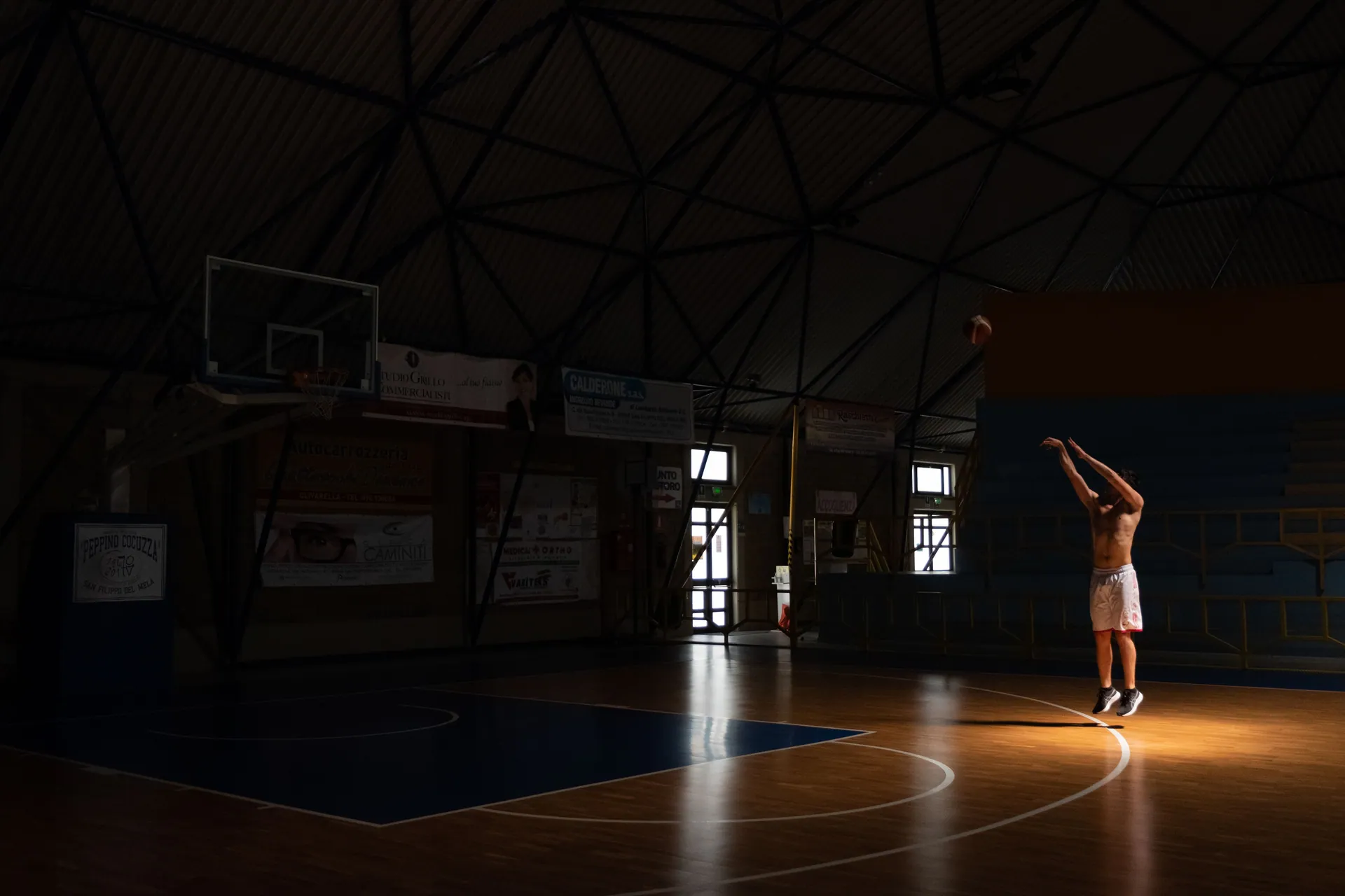 A man practices basketball shots in a dimly lit indoor court, creating a dramatic and focused atmosphere.