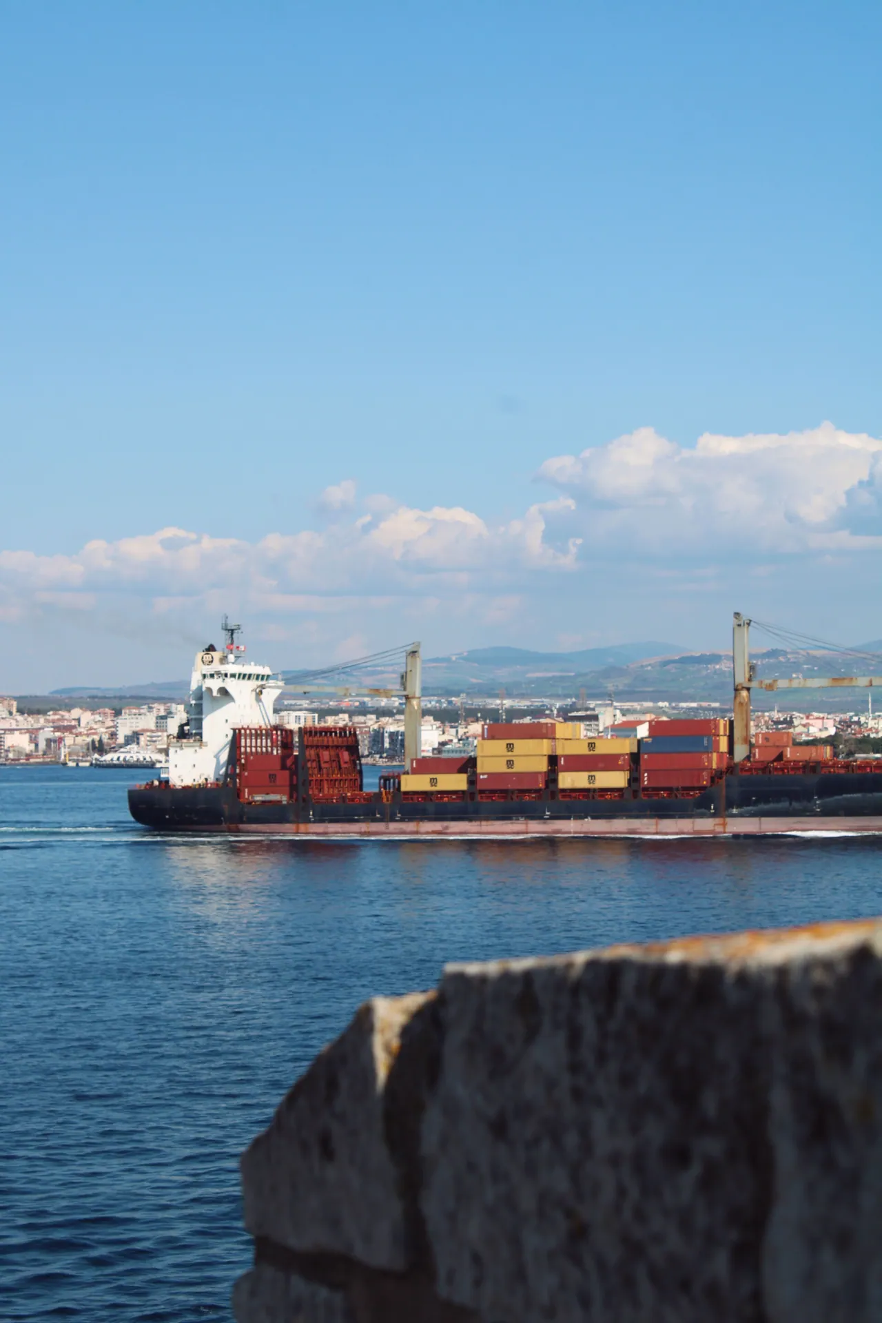 A cargo ship loaded with containers sails past a coastal city with clear blue skies.