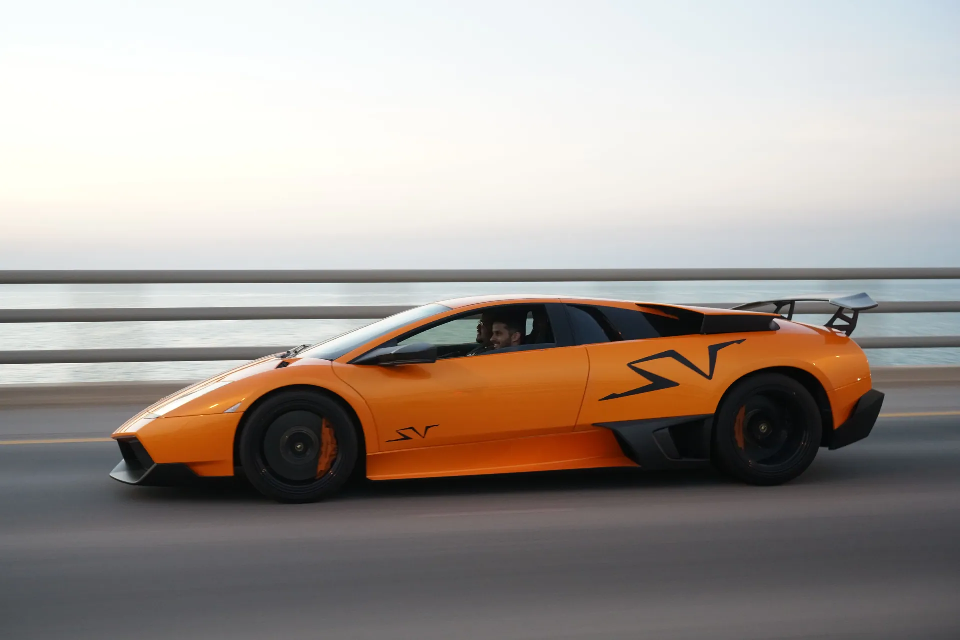 A vibrant orange sports car drives along a coastal road, showcasing luxury and speed.