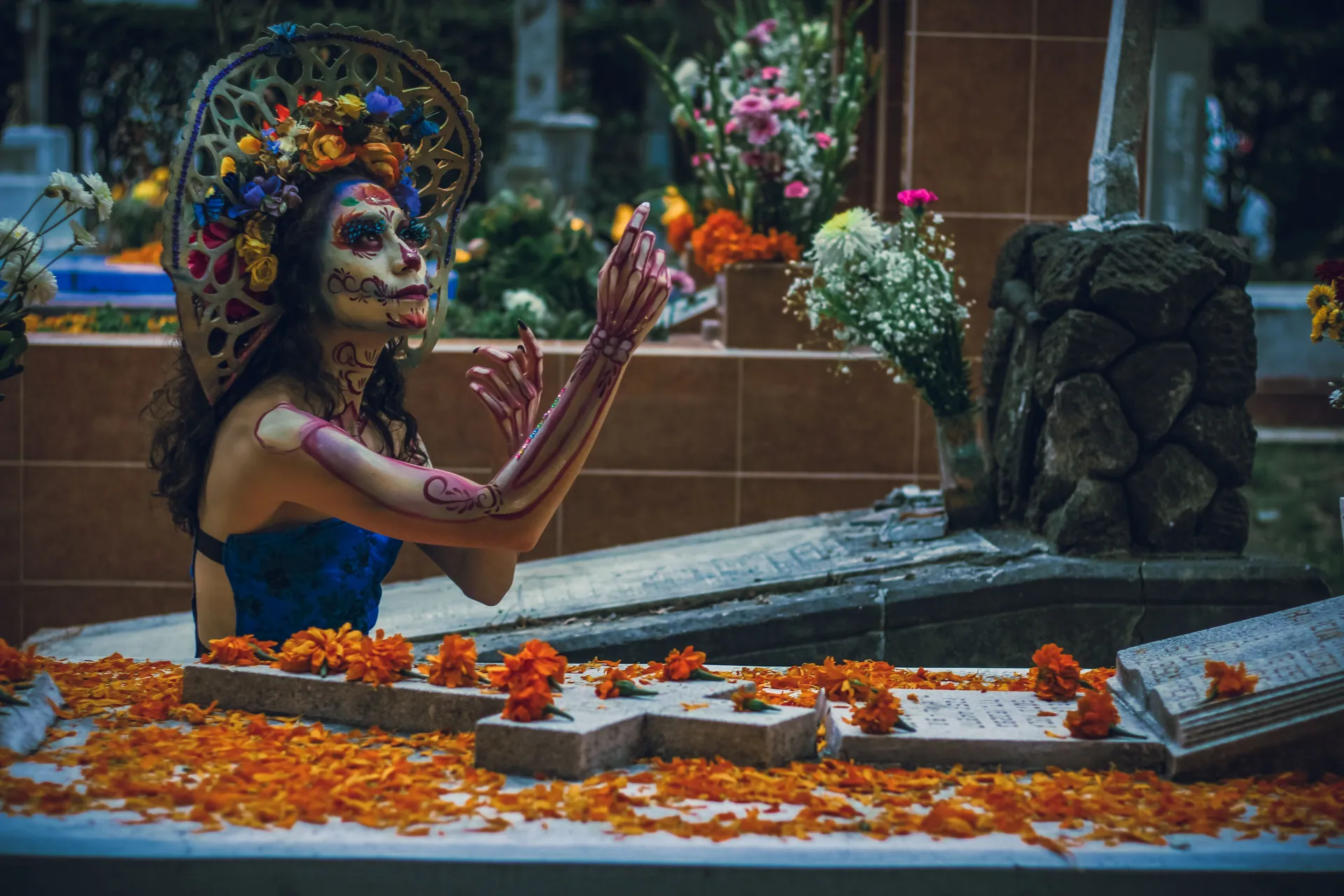A woman in vibrant Dia de Muertos attire at a flower-adorned grave.