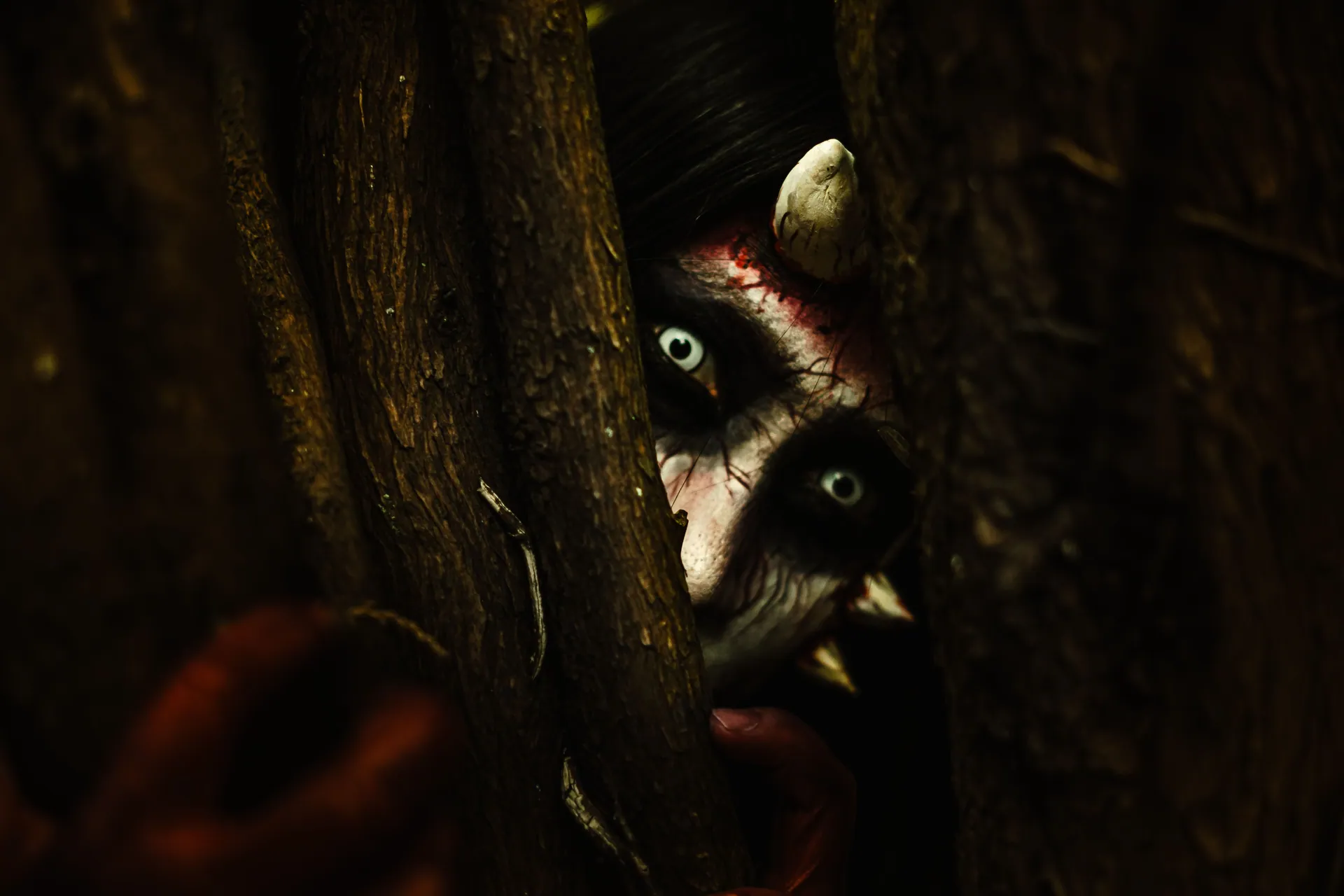 A person with devilish makeup peers through tree branches, creating an eerie Halloween vibe.