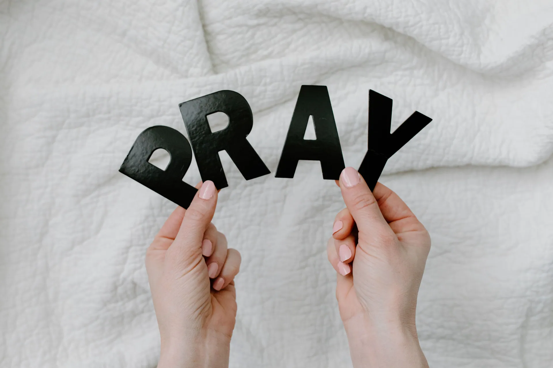 Close-up of hands holding black cutout letters spelling PRAY on white fabric background.