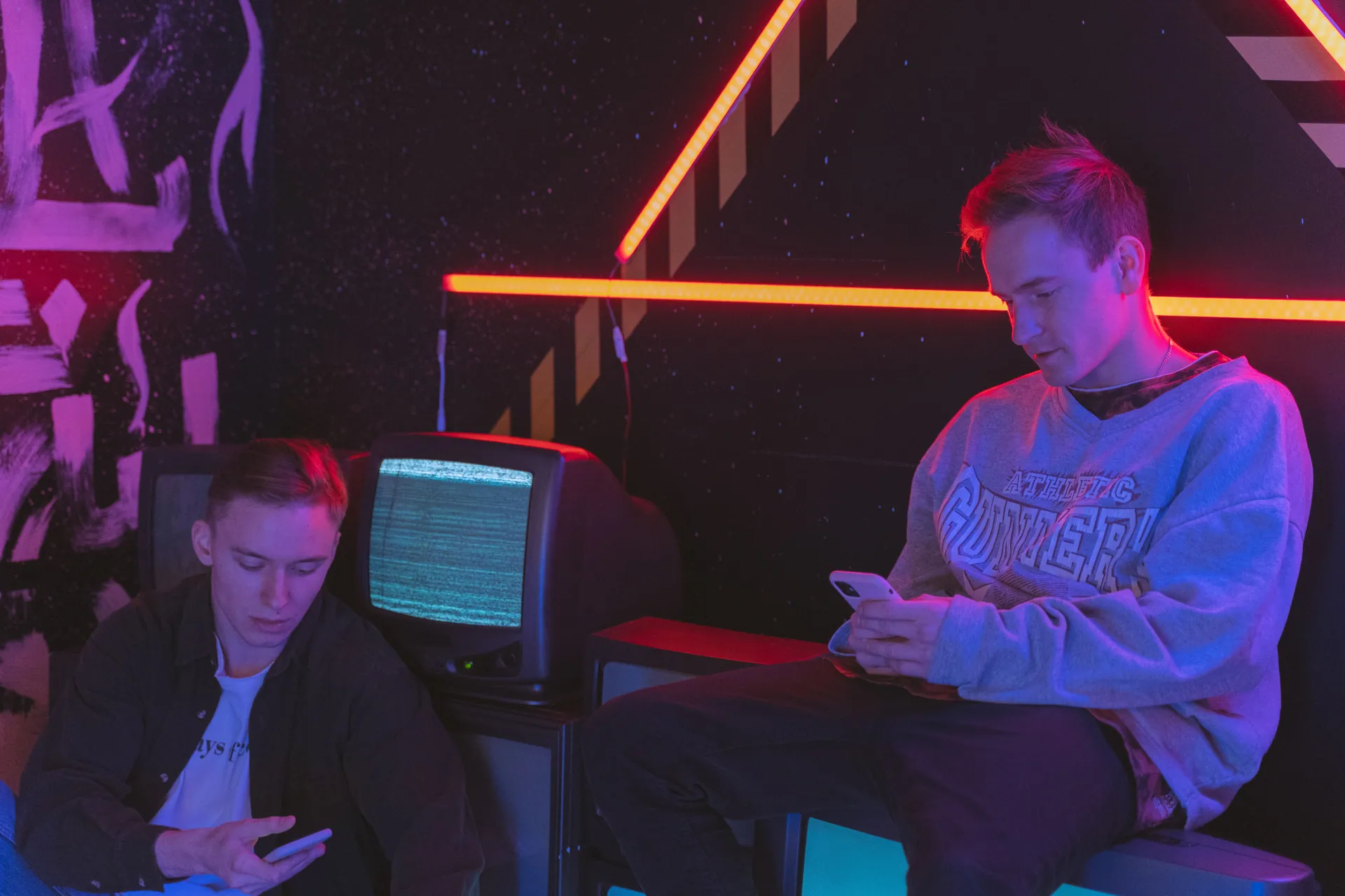 Two young men using smartphones in a neon-lit room with vintage TV and urban graffiti style.