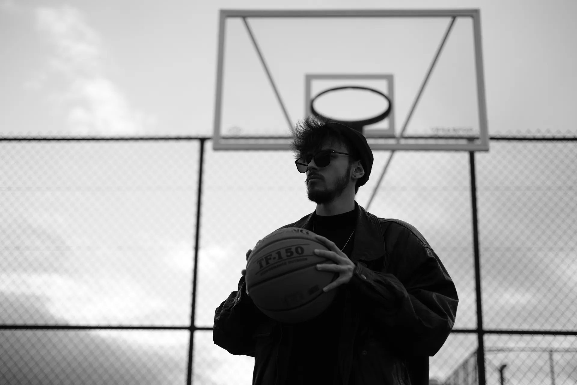 Black and white photo of a young man posing with a basketball outdoors on a court.