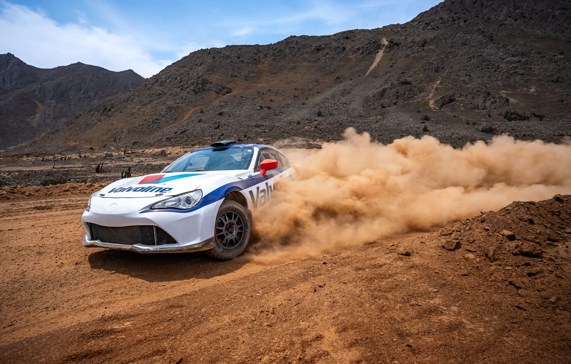 Rally car drifts on dusty, mountainous dirt track, kicking up dust clouds.