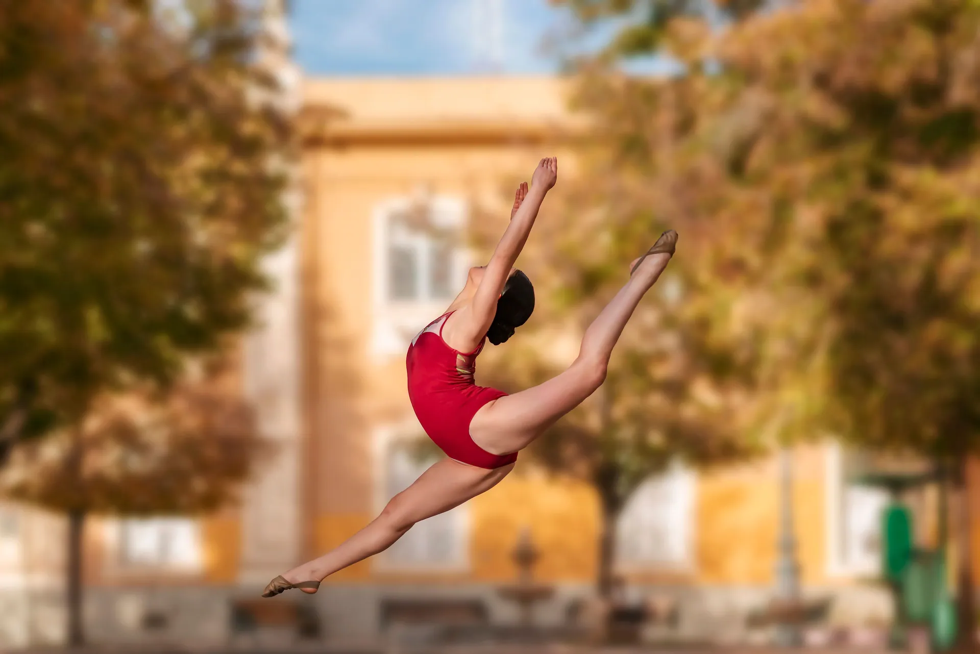 Aerial view of a female dancer in a red leotard performing a mid-air split outdoors.
