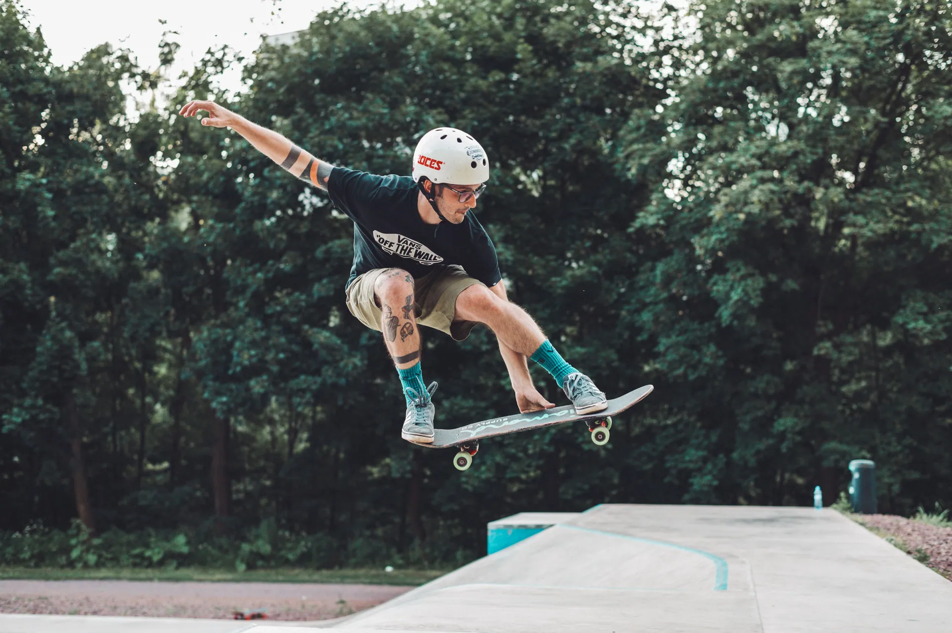 Young man performing a skateboard trick at an outdoor skatepark surrounded by trees.
