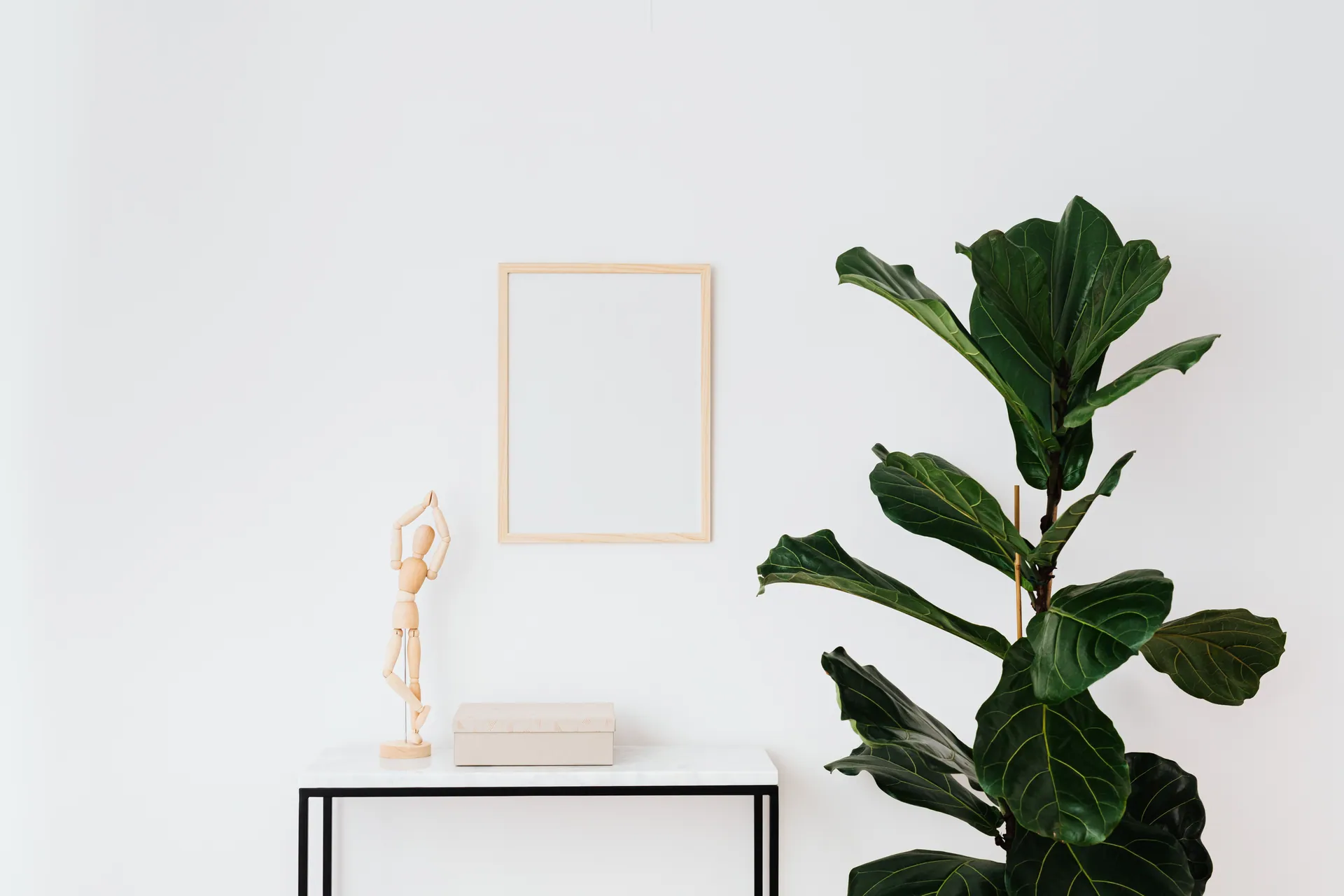 Simple interior with fiddle leaf fig, wooden figure, and white frame on a desk.