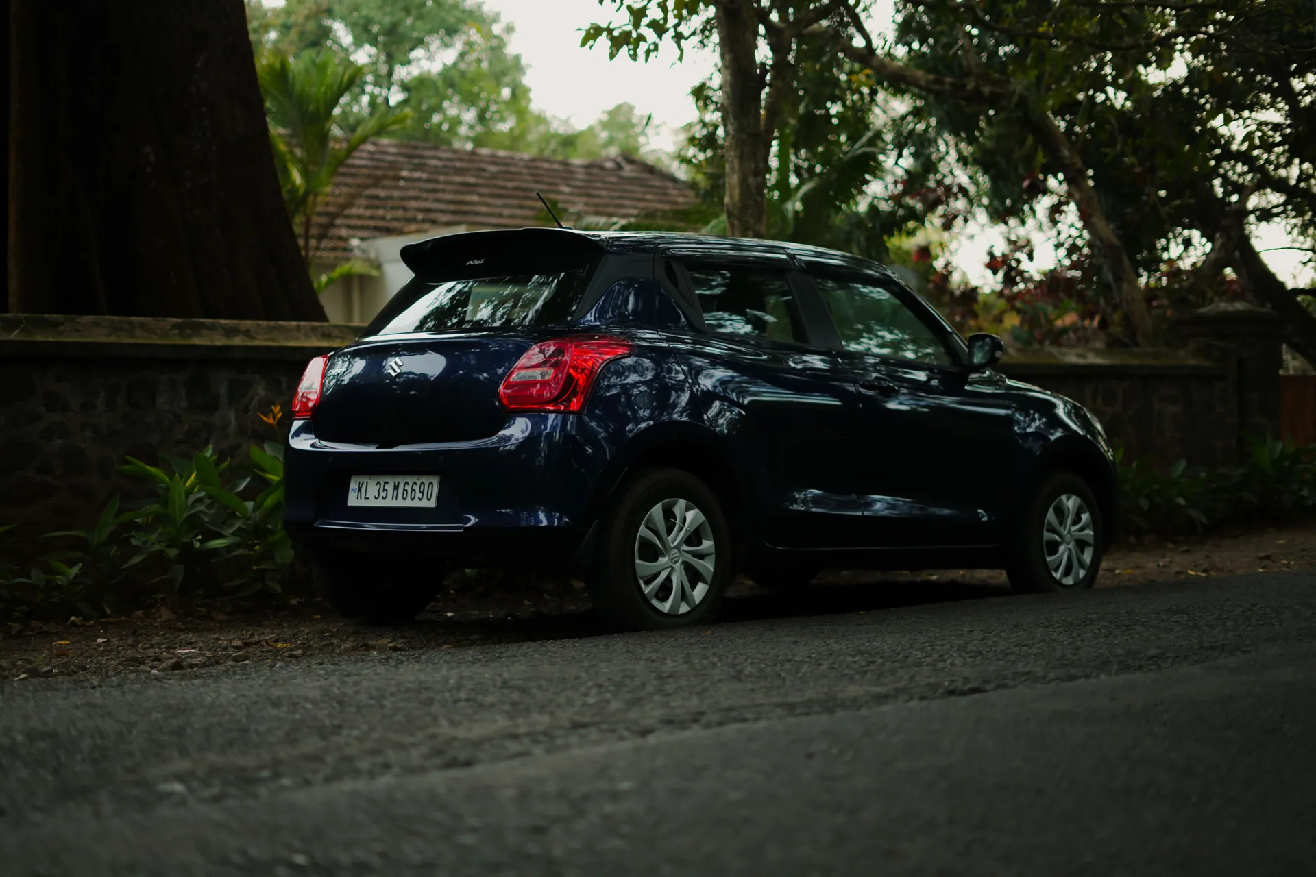 A blue compact car parked on a quiet street with lush greenery and rustic architecture in India.