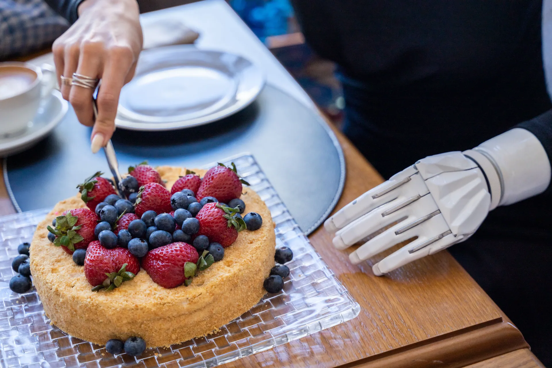A person with a prosthetic arm cuts a cake topped with strawberries and blueberries, showcasing modern technology.