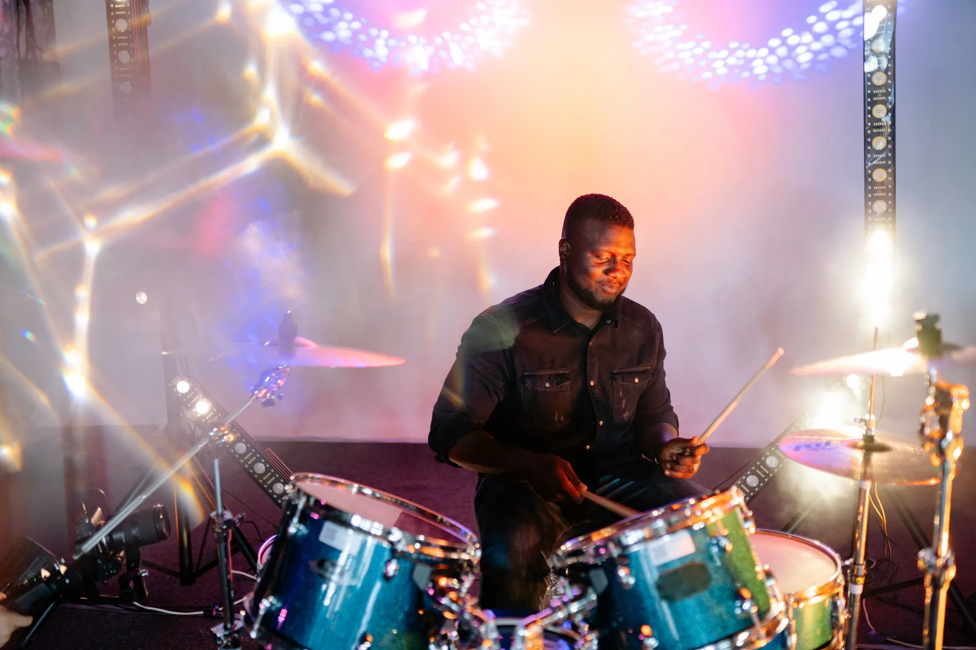 African American drummer playing energetically under colorful stage lights.
