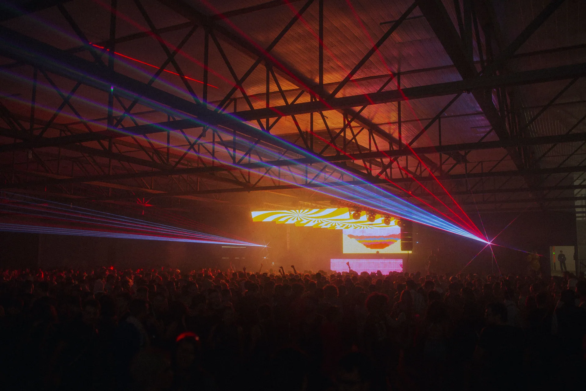 A lively indoor concert featuring colorful laser lights over a large crowd enjoying music.
