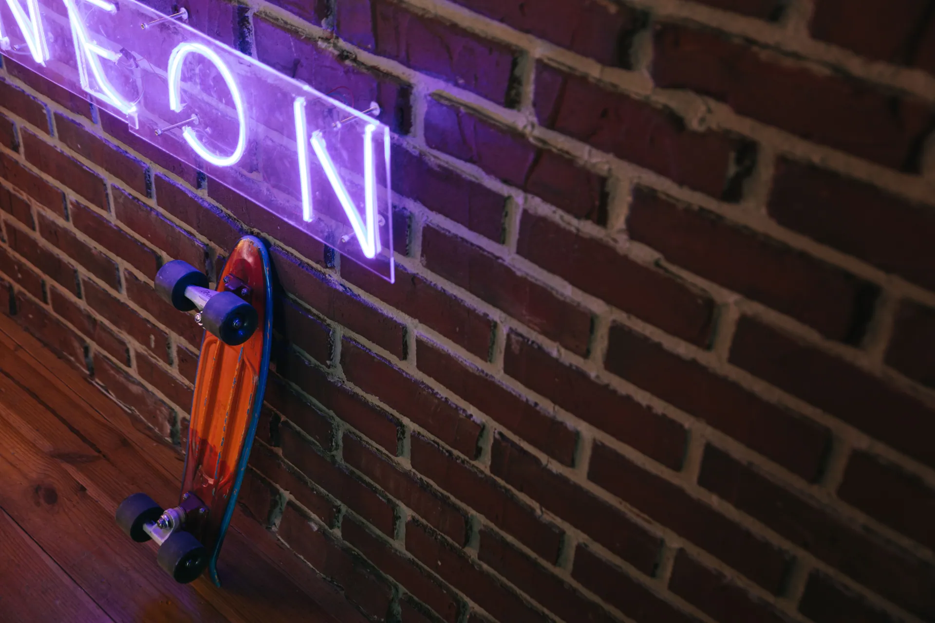 A neon sign glows above a penny board resting on a rustic brick wall indoors.