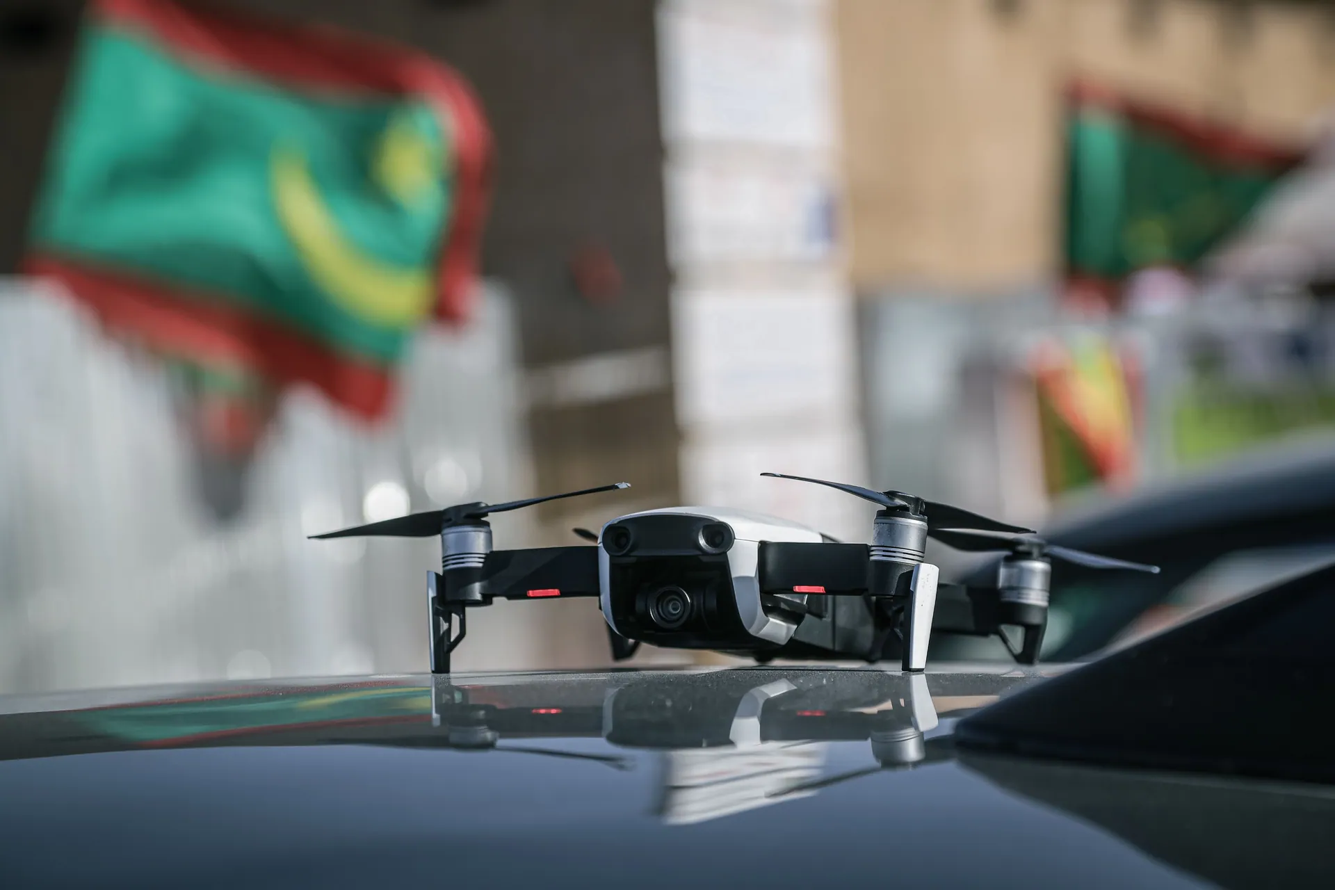 A small drone rests on a car hood, reflected with flags in the background.