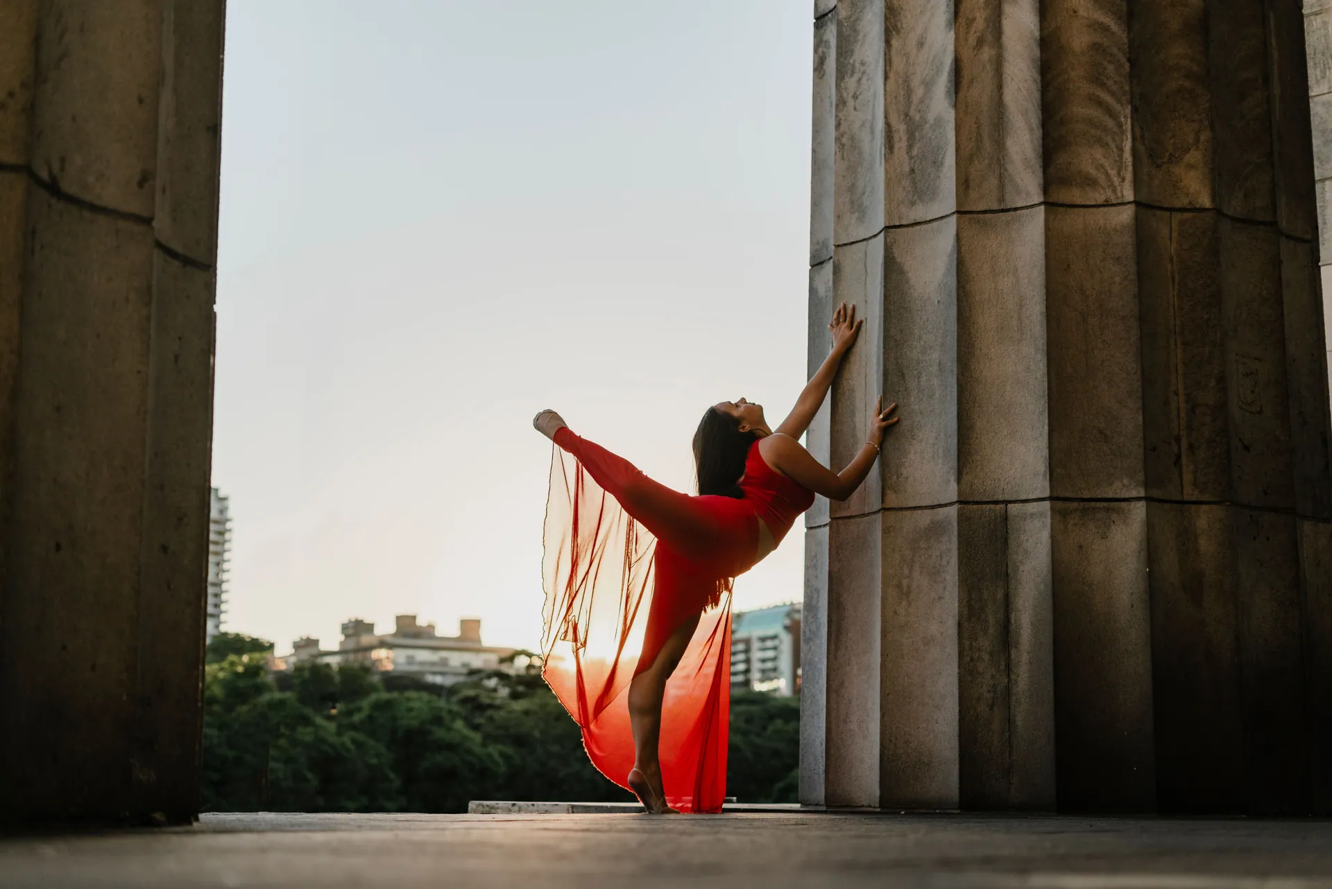 A graceful female dancer in a red dress striking a pose between city pillars during dusk.