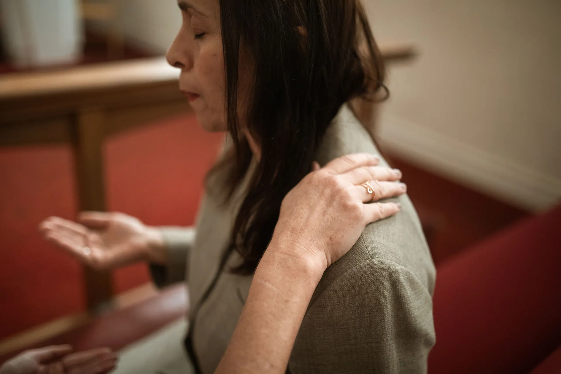 A woman prays indoors, with another person's hand on her shoulder for comfort, expressing faith and spirituality.