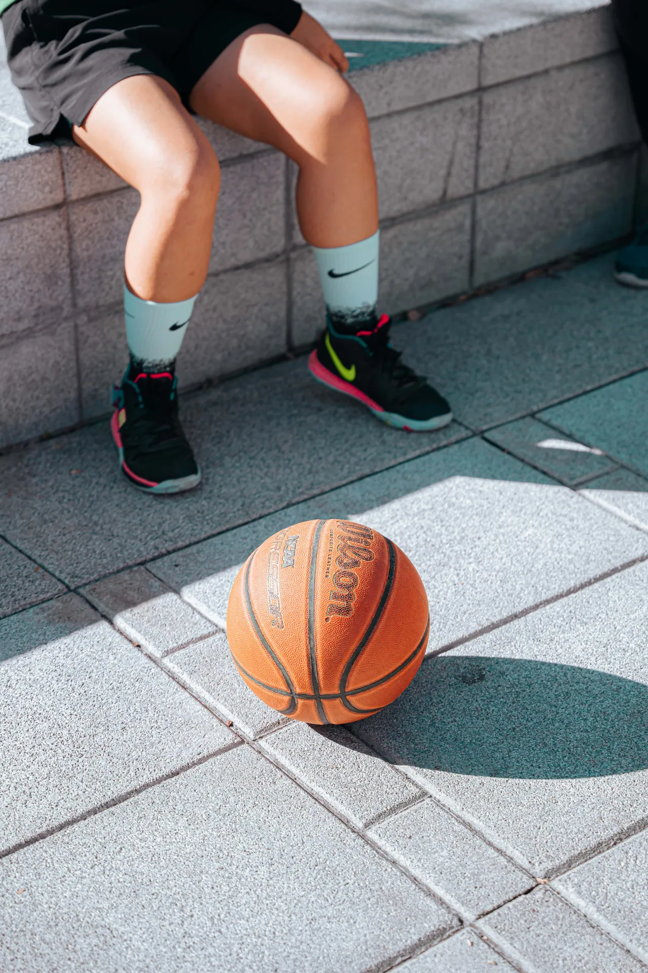 A basketball rests outdoors on a sunny day with a player's legs in the background.