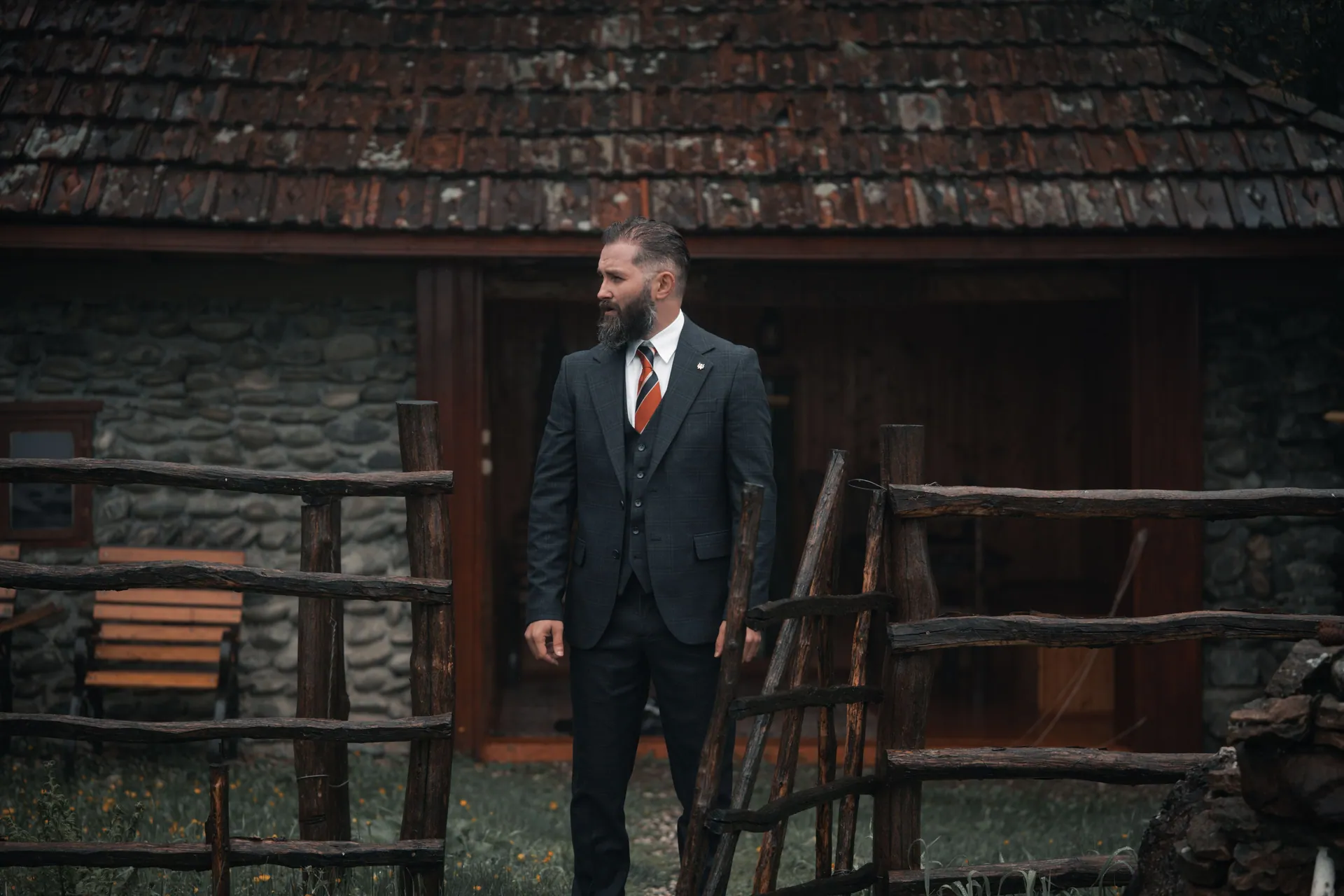 Bearded man in a formal suit standing outside a rustic stone cottage with wooden fence.