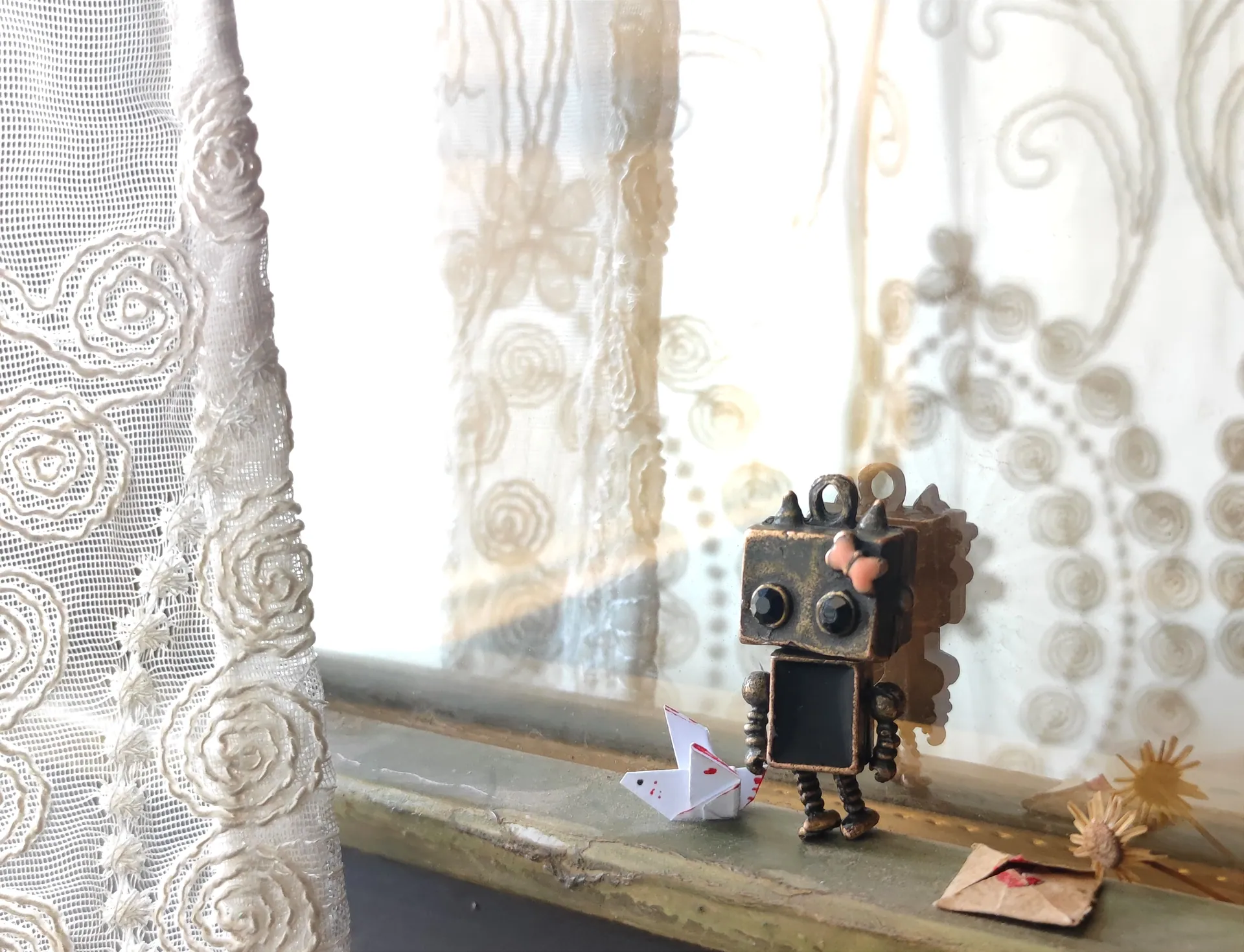 A charming robot figurine with an origami heart sits on a windowsill next to lace curtains, evoking a playful scene.